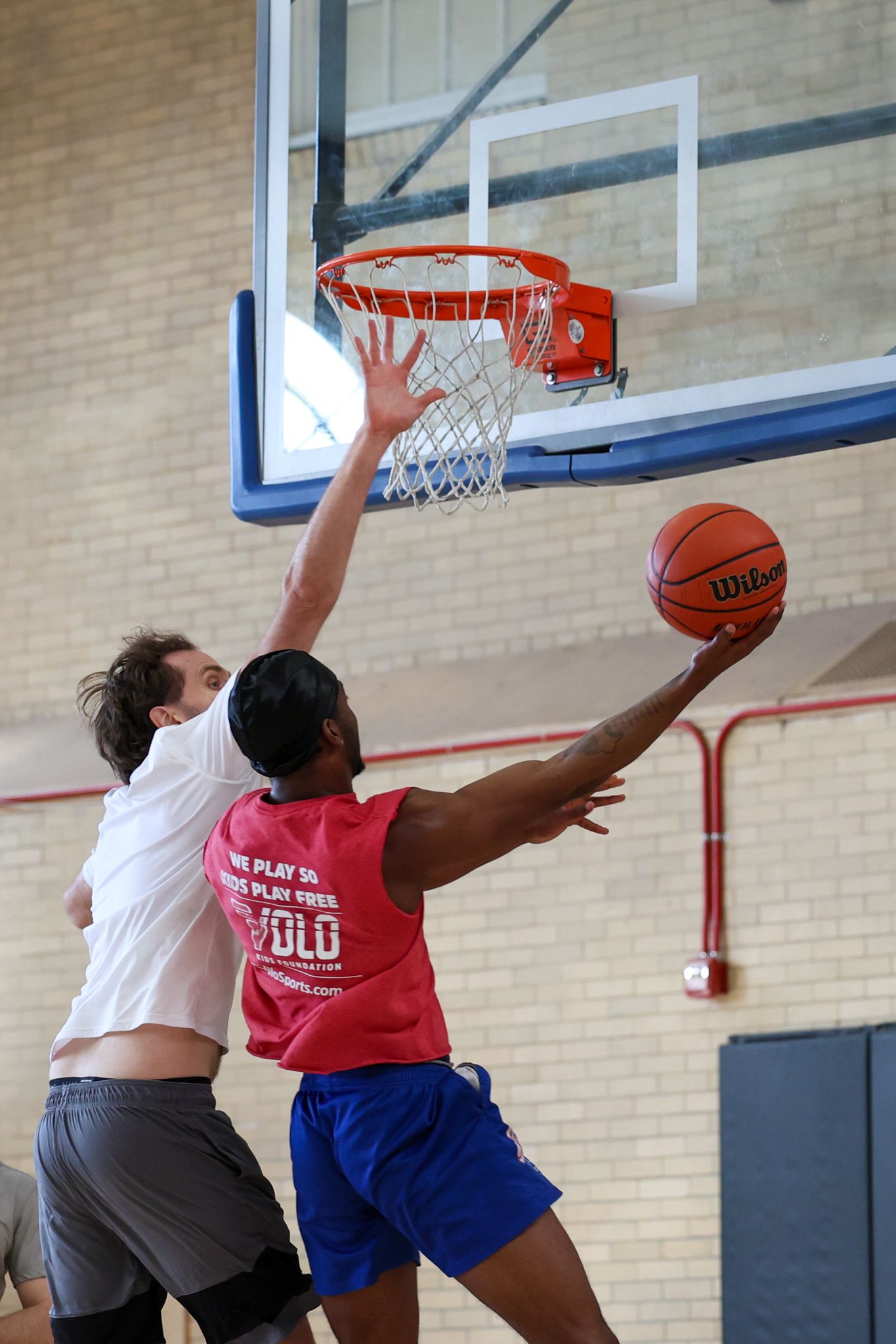 Two men play basketball indoors, one attempting a layup while the other blocks.