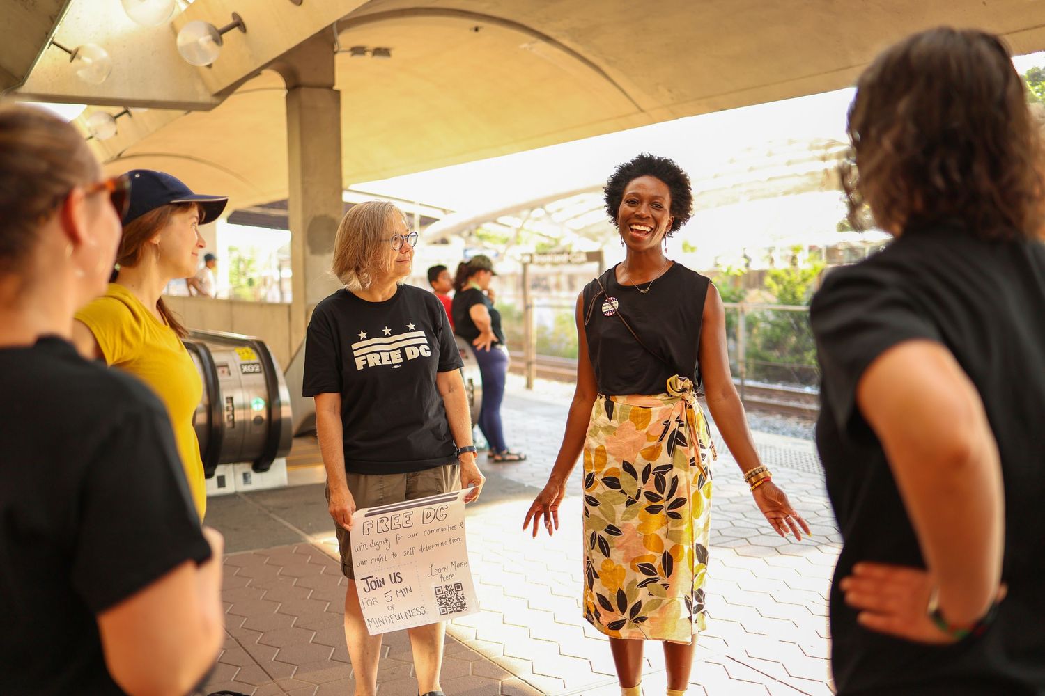 Smiling Black woman in floral skirt speaks to a diverse group of activists at an outdoor transit station.