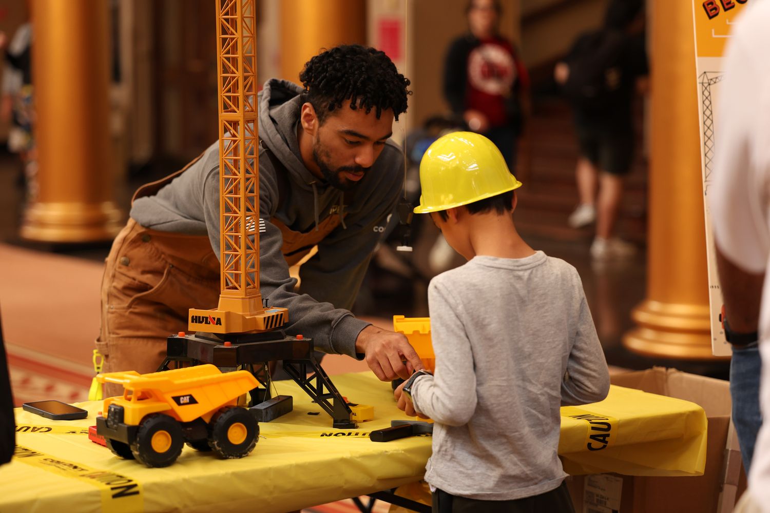 Man helps child with toy construction vehicles at a yellow table during an indoor educational event.