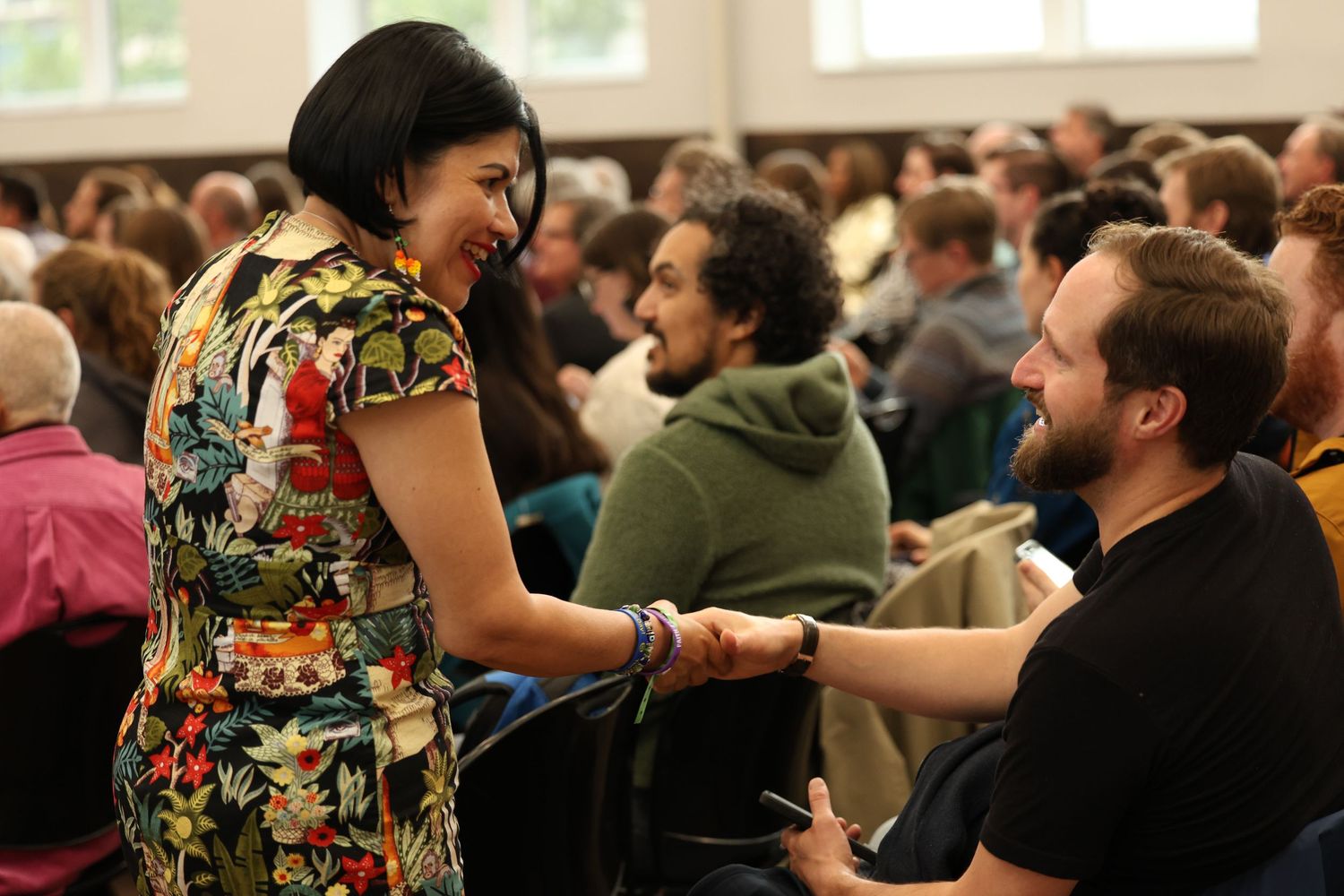 Woman in floral dress shakes hands with bearded man at a crowded indoor gathering.