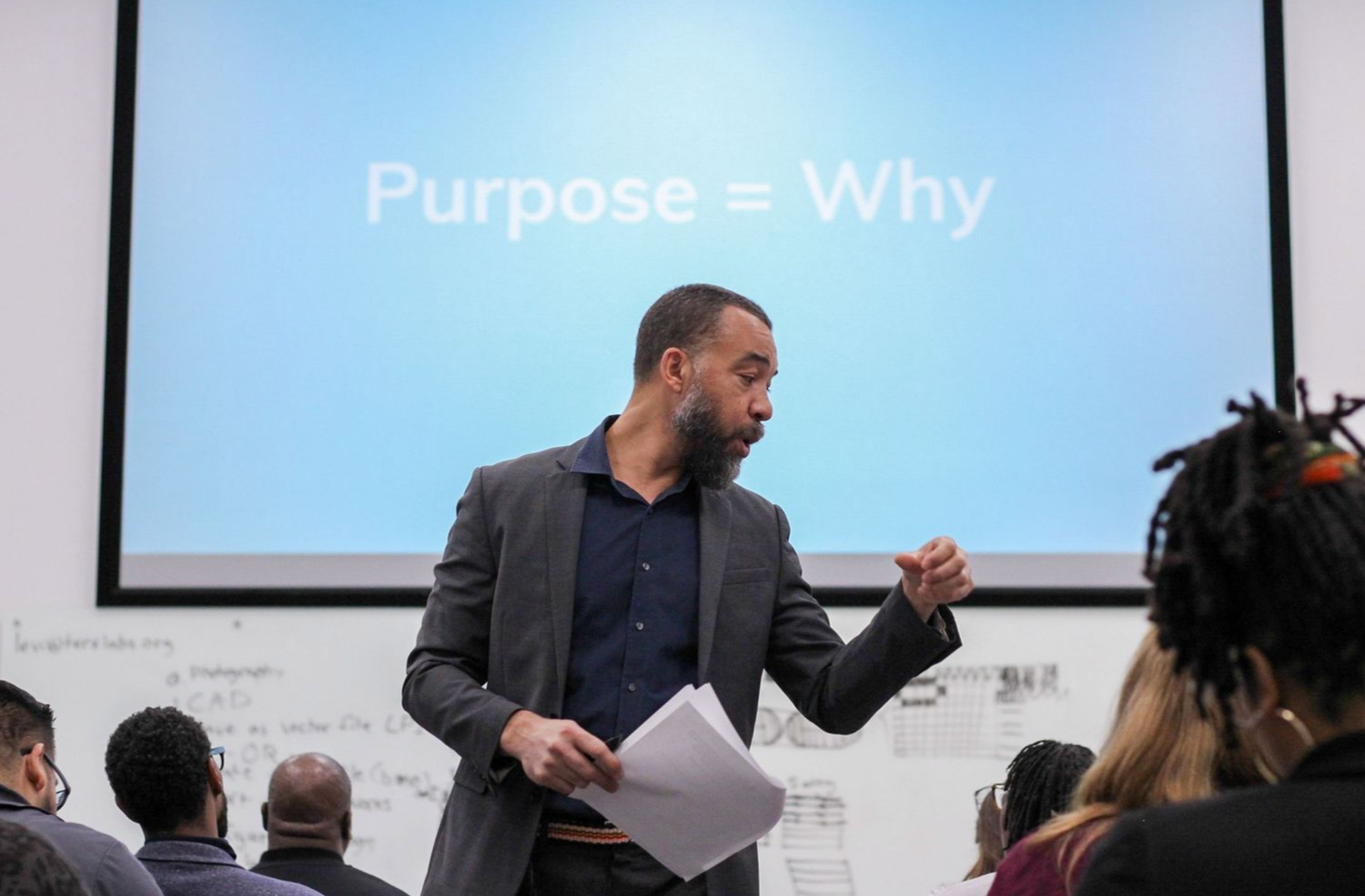 A speaker presents in front of a screen displaying Purpose equals Why during a professional workshop.