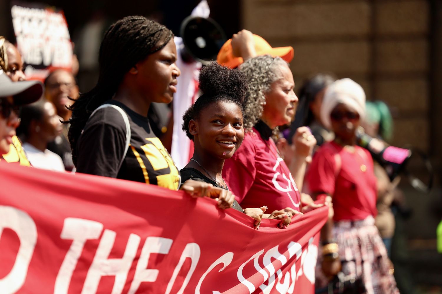 Diverse group of women marching in protest, holding a red banner at a civil rights demonstration outdoors.