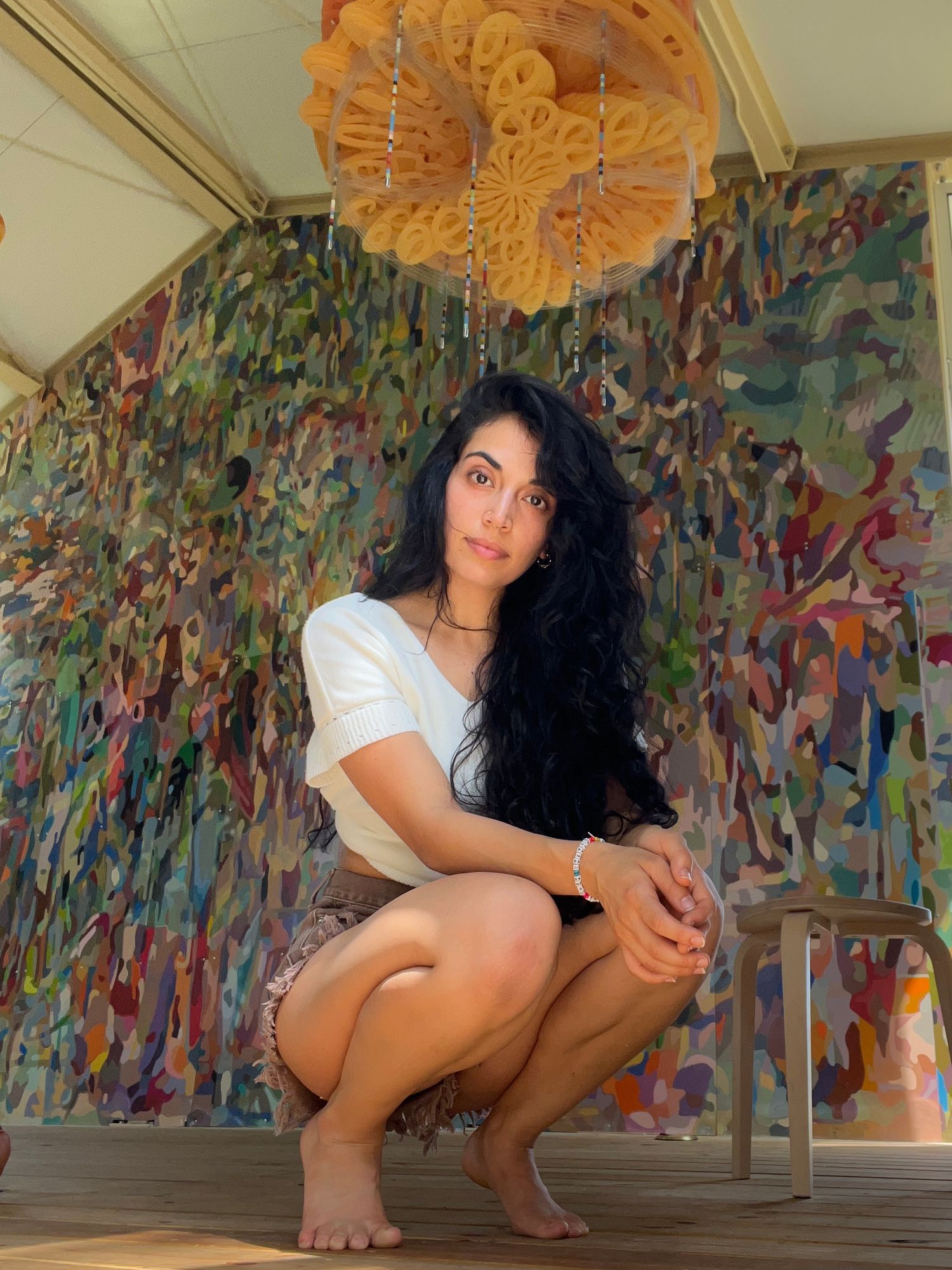 Young woman with long dark curly hair crouching barefoot in front of a colorful abstract mural wall.