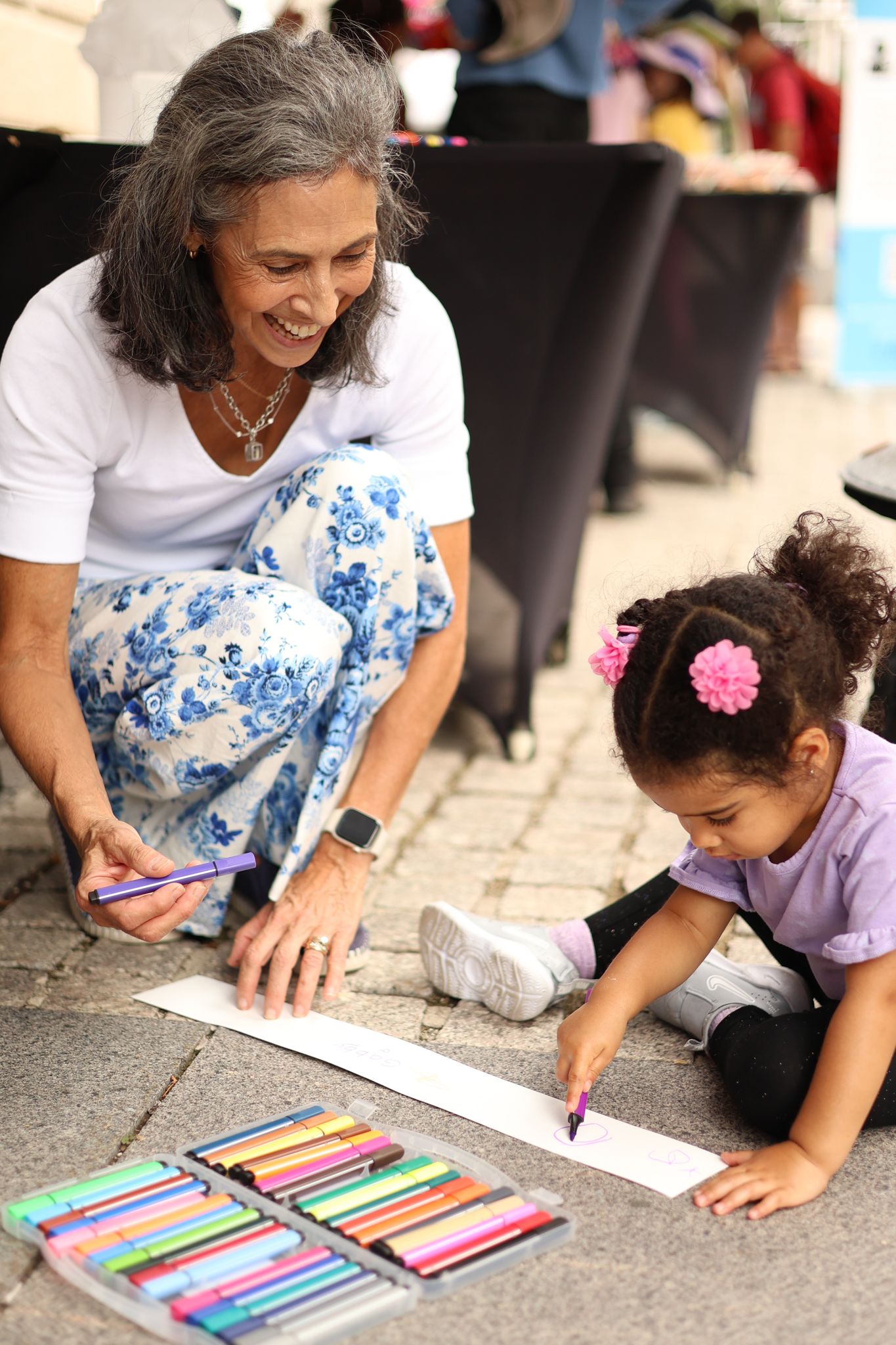 A woman smiles while helping a young girl draw with colorful markers on paper outdoors.