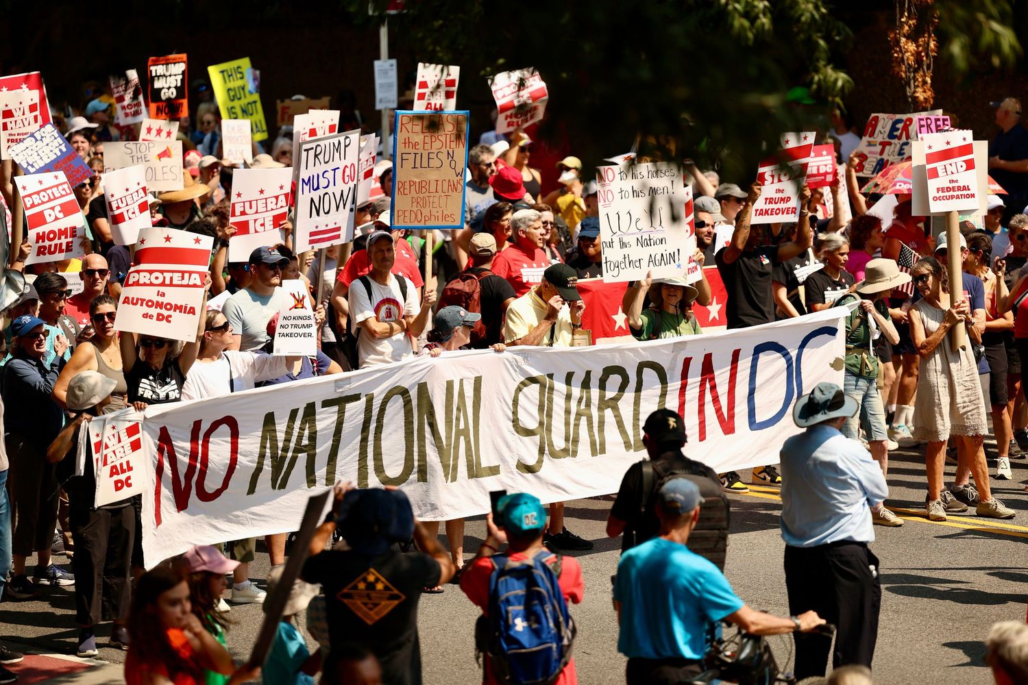 Large crowd of protesters carrying signs march behind a banner reading No National Guard In DC.