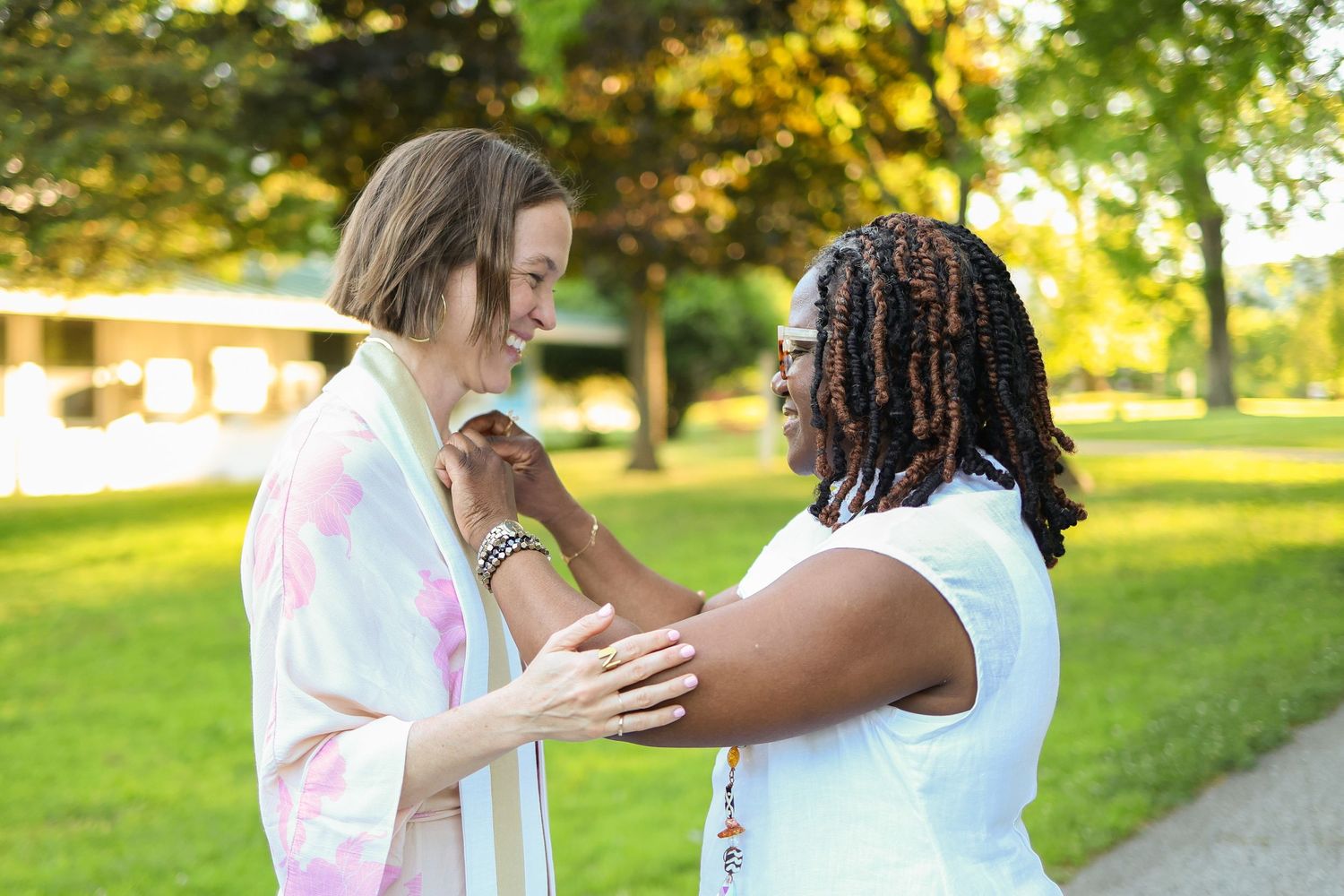 Two women smiling and holding hands in a sunny park, sharing a warm and joyful moment outdoors.