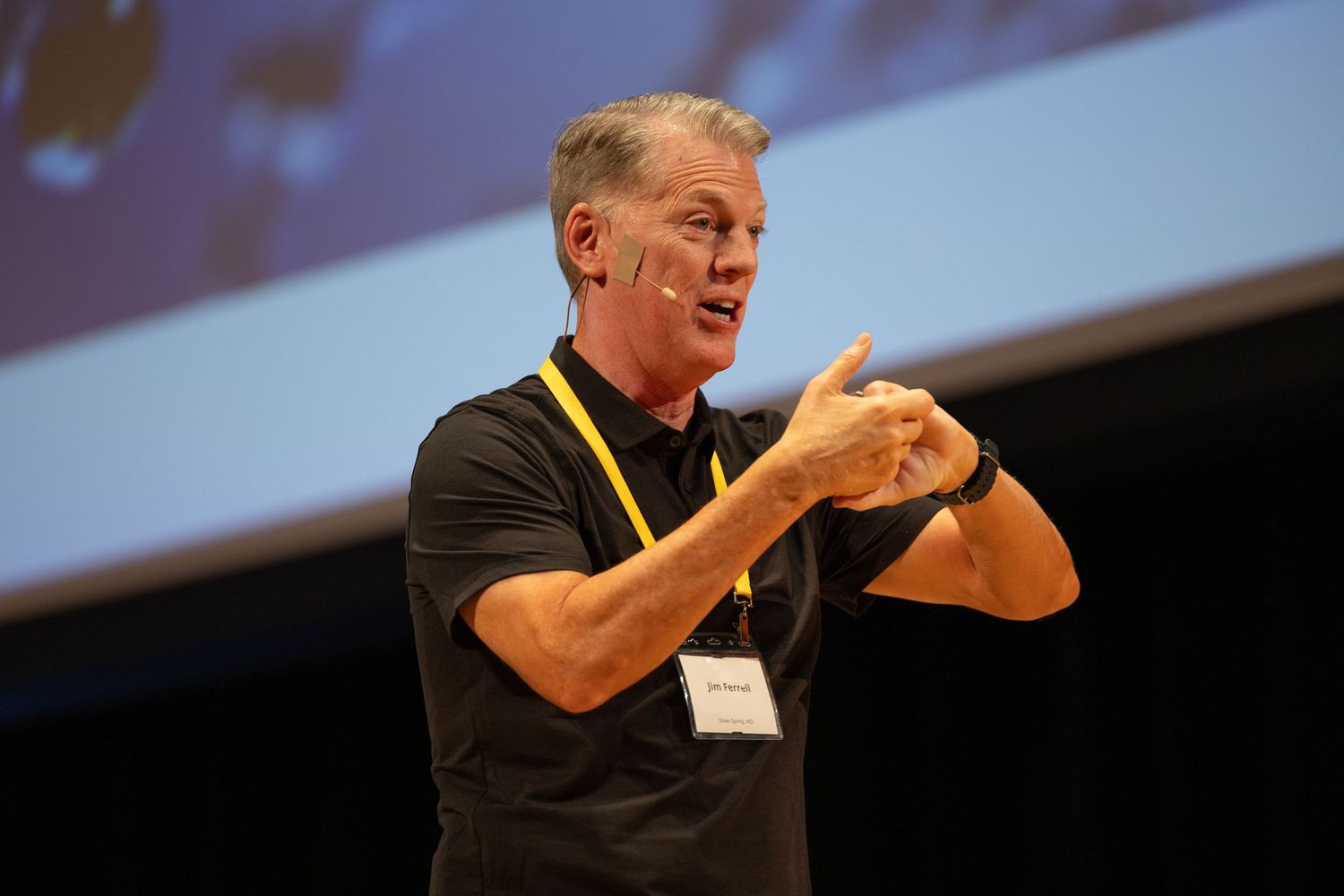 A male speaker gestures on stage wearing a black polo and yellow lanyard during a conference presentation.
