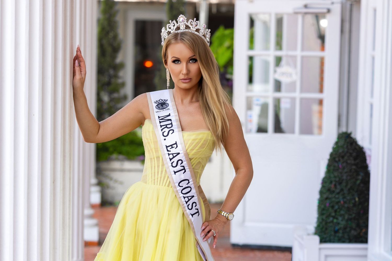 Mrs. East Coast titleholder in a yellow strapless gown, tiara, and sash leaning against a white architectural pillar