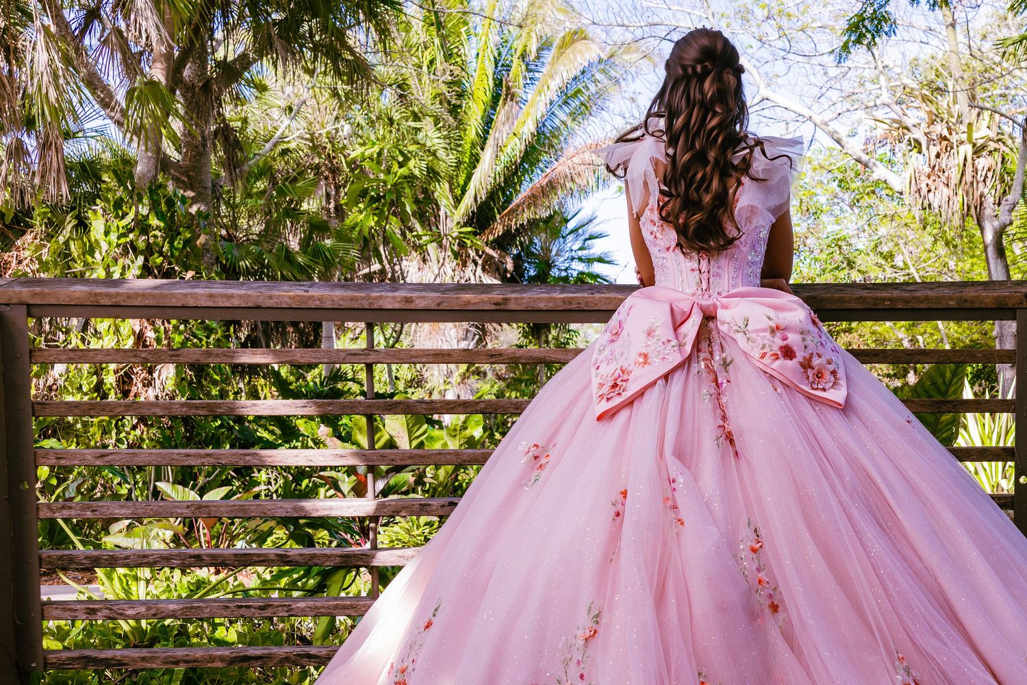 Back view of a young woman with long wavy brown hair wearing a pink embroidered ballgown next to a balcony railing