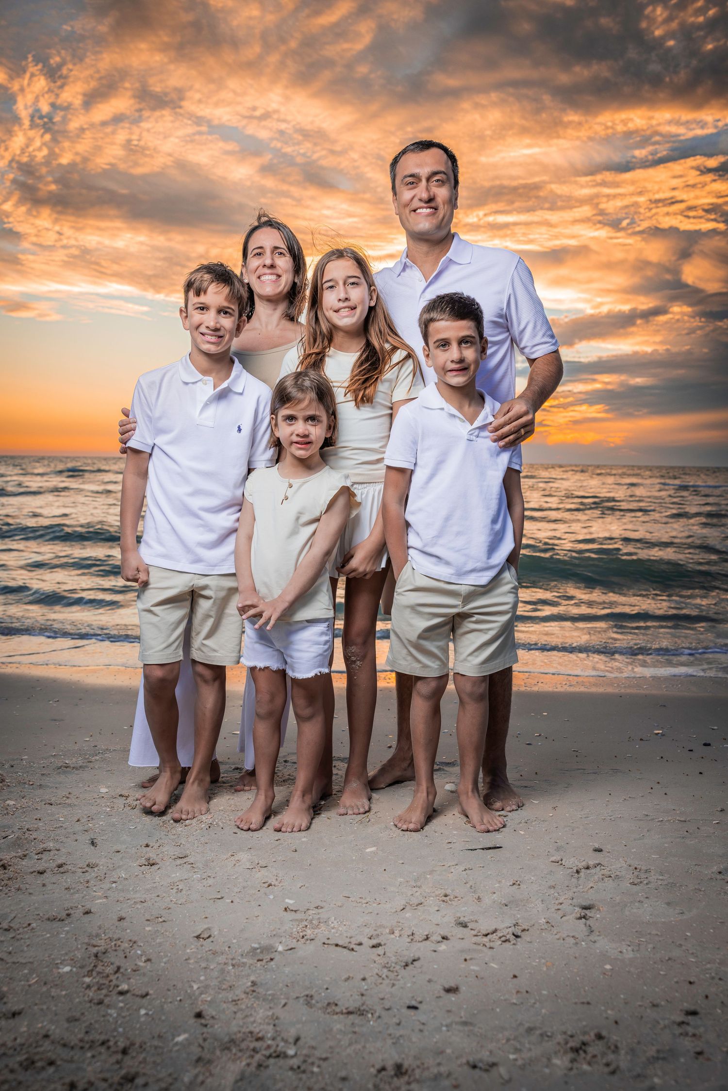 Family of six with young children standing together on a sandy beach during a vibrant orange sunset.
