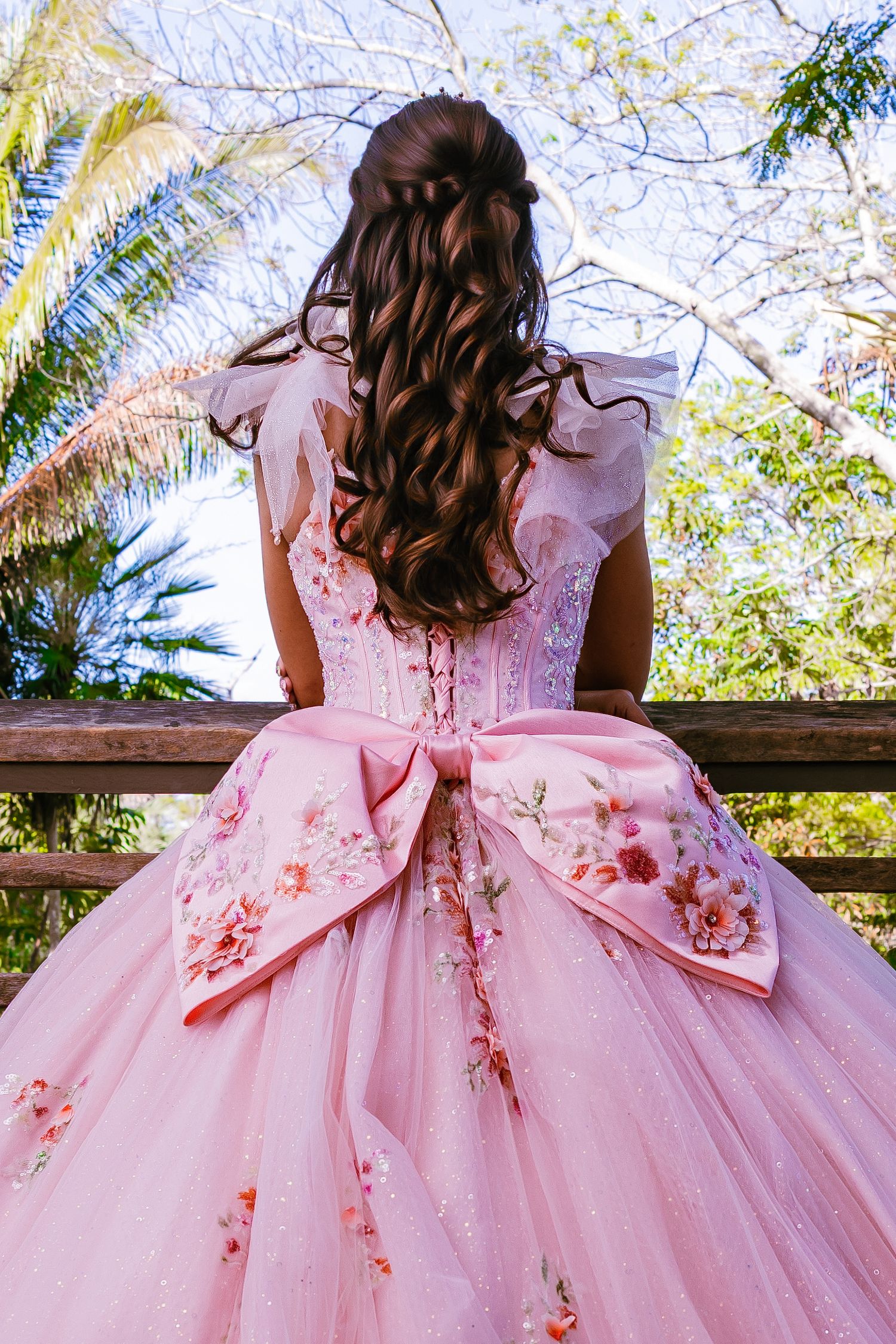 Back view of a young woman with long wavy brown hair wearing a pink embroidered ballgown