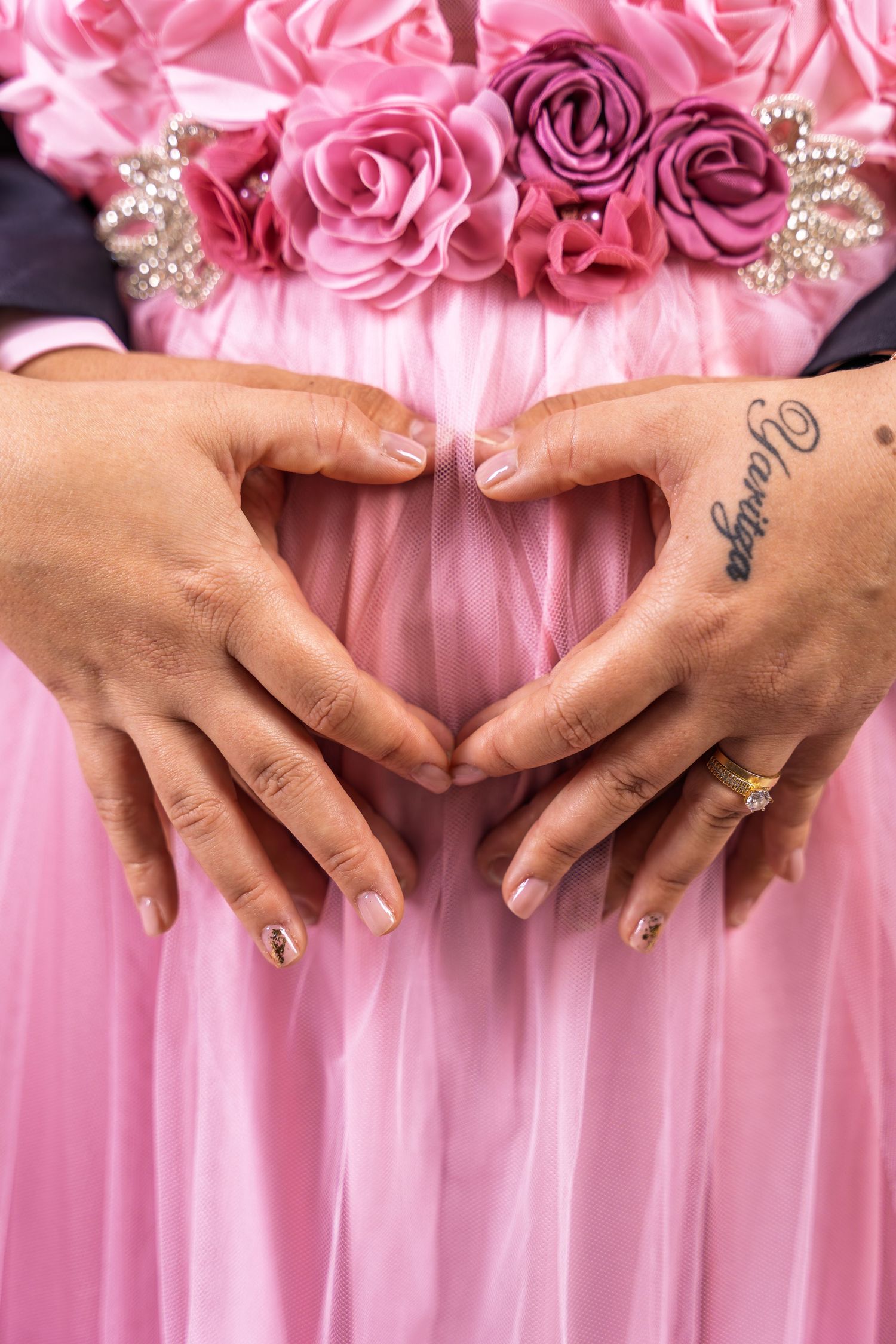 Close-up of hands with tattoos forming a heart shape over a pregnant belly in a light pink dress adorned with fabric flowers.