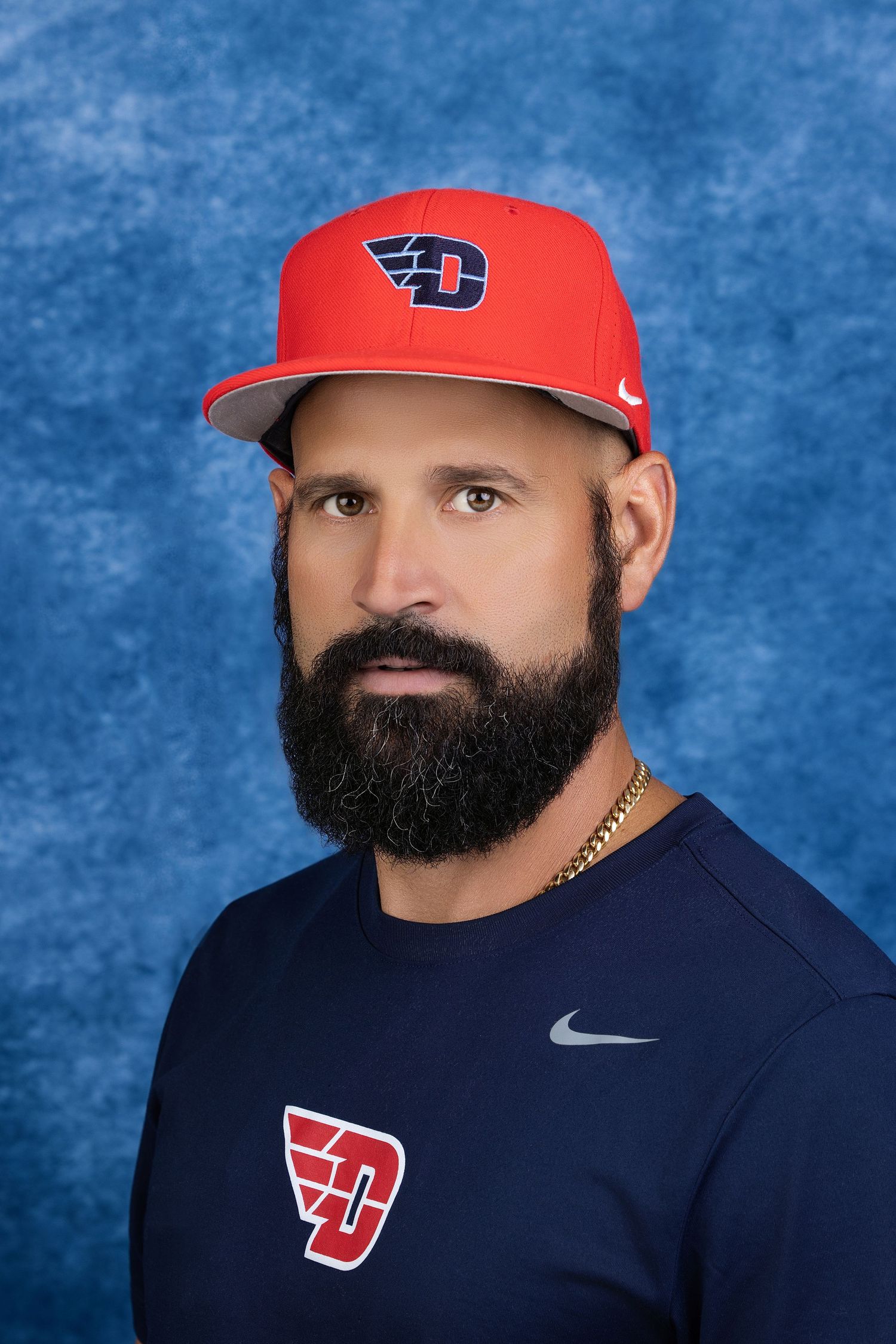 Bearded man wearing a red baseball cap and navy blue shirt against a blue background
