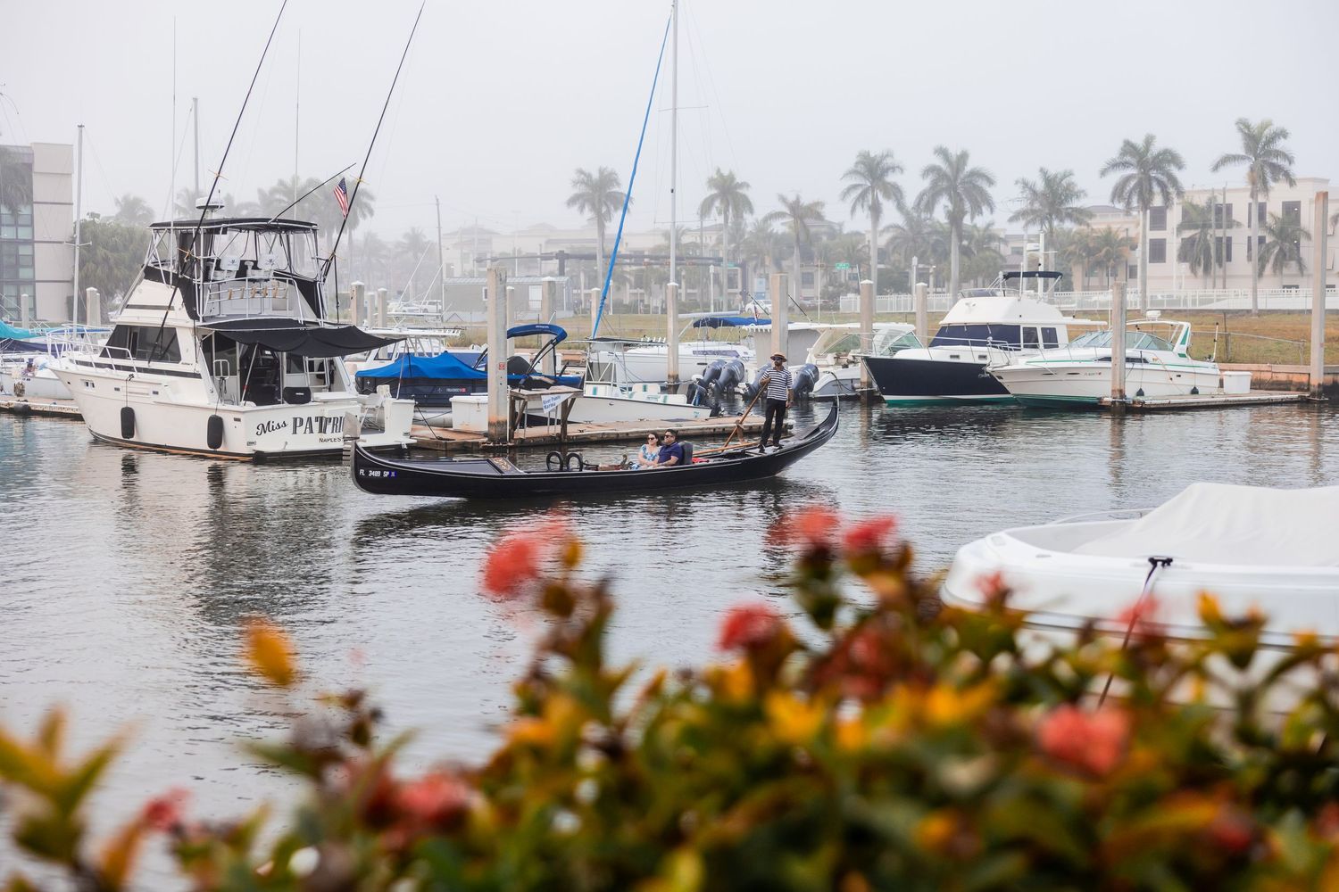 Traditional gondola cruising past docked boats in a foggy marina behind colorful foreground flowers