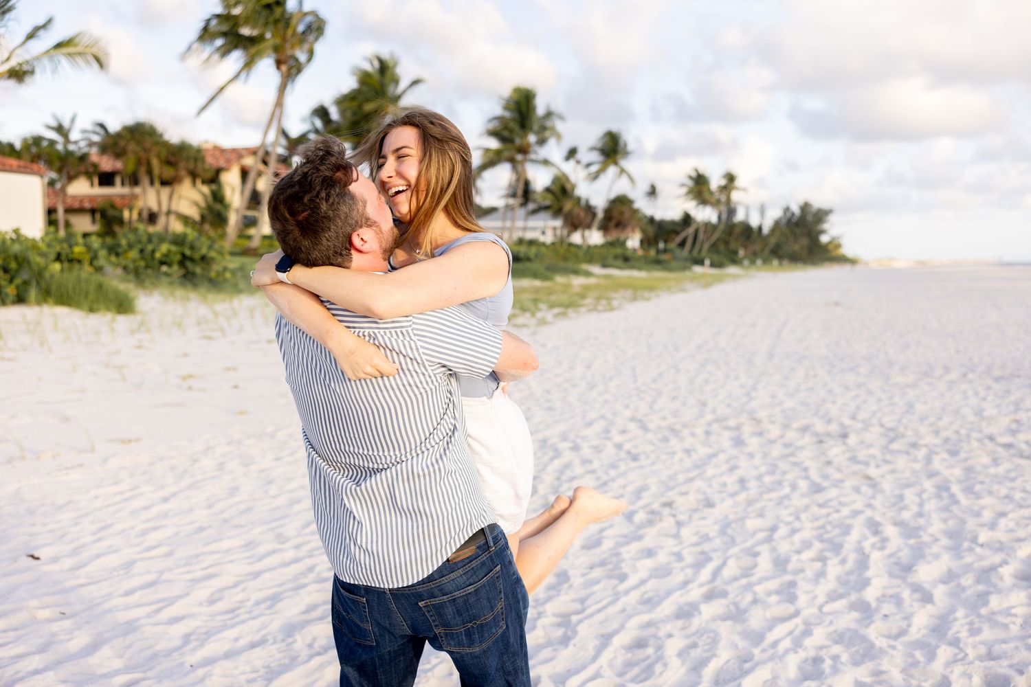 Man lifting and hugging a laughing woman on a white sand beach with palm trees and ocean background
