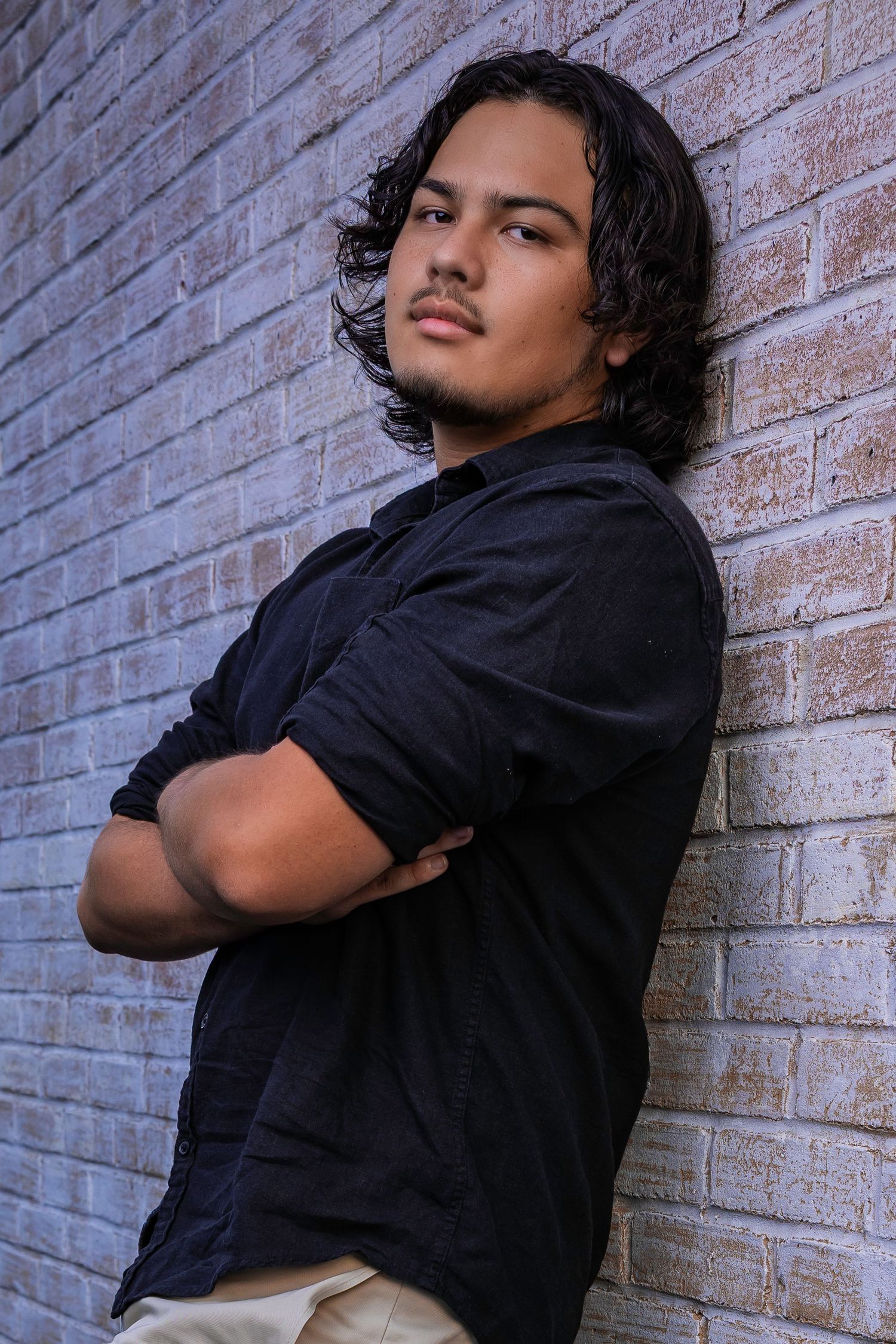 Young man with wavy shoulder-length hair standing with arms crossed against a brick wall.