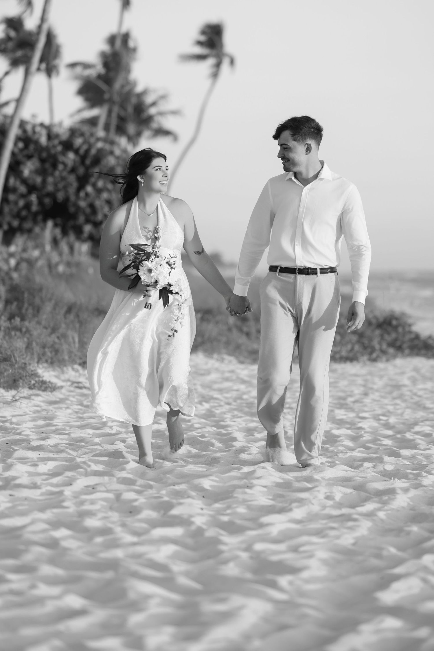 Black and white photo of a bride and groom walking hand-in-hand along a sandy beach