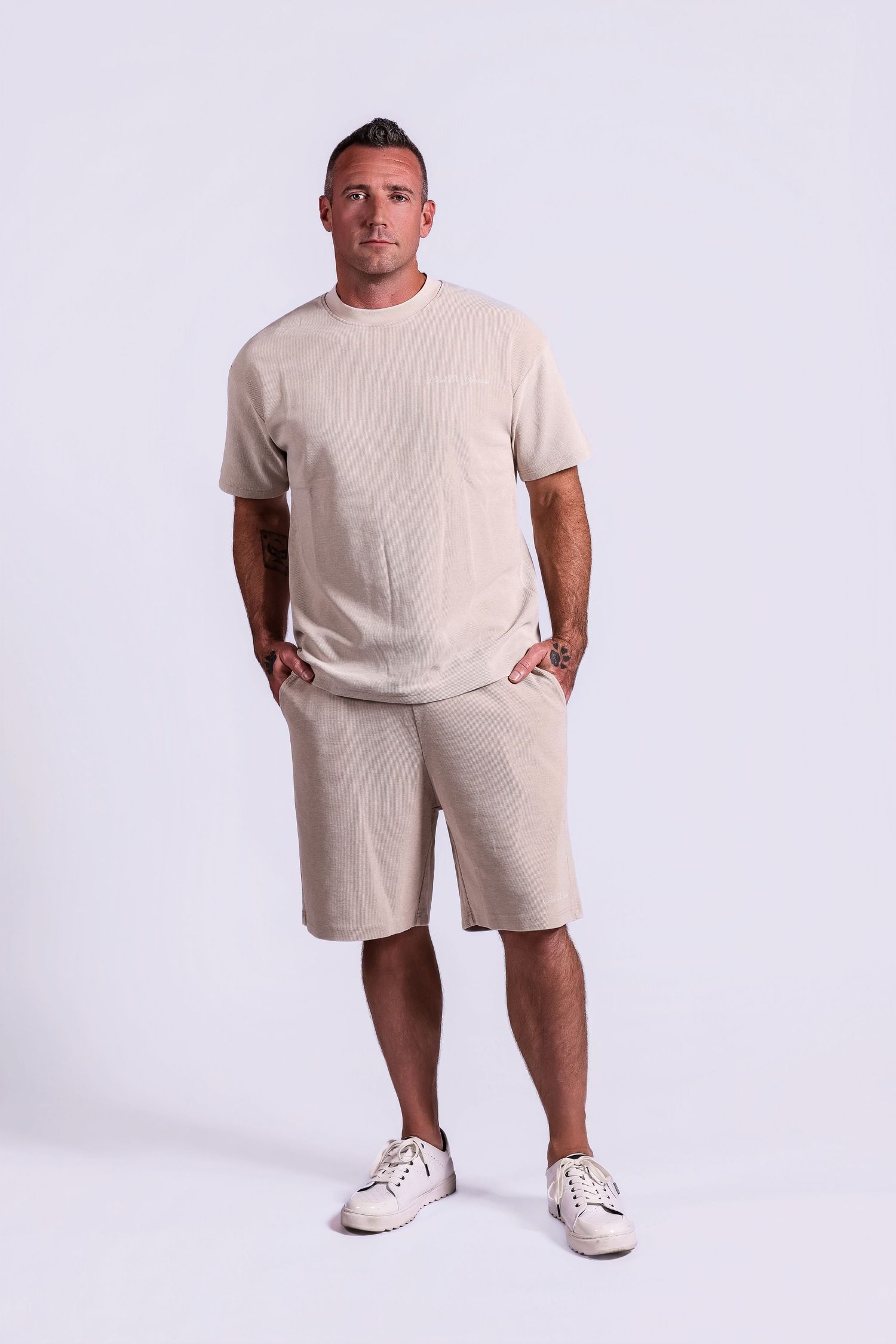 Full-length studio portrait of a man standing with hands in pockets wearing a loose-fitting cream t-shirt, matching shorts, and white sneake