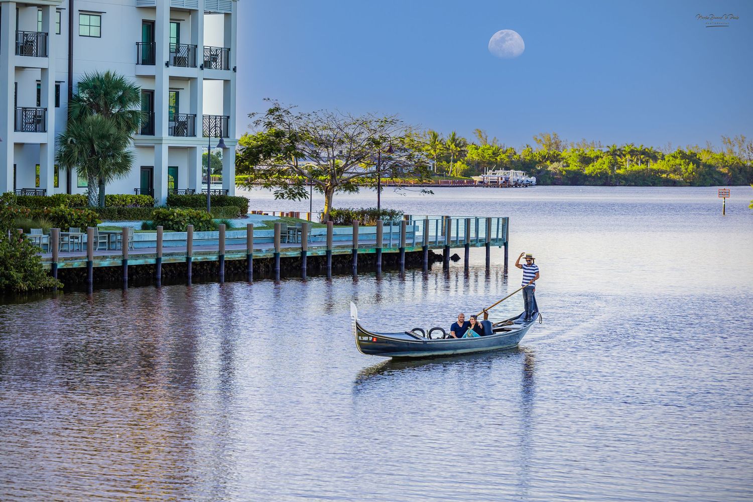 Gondolier rowing a classic gondola boat across a calm river with waterfront properties in the background.