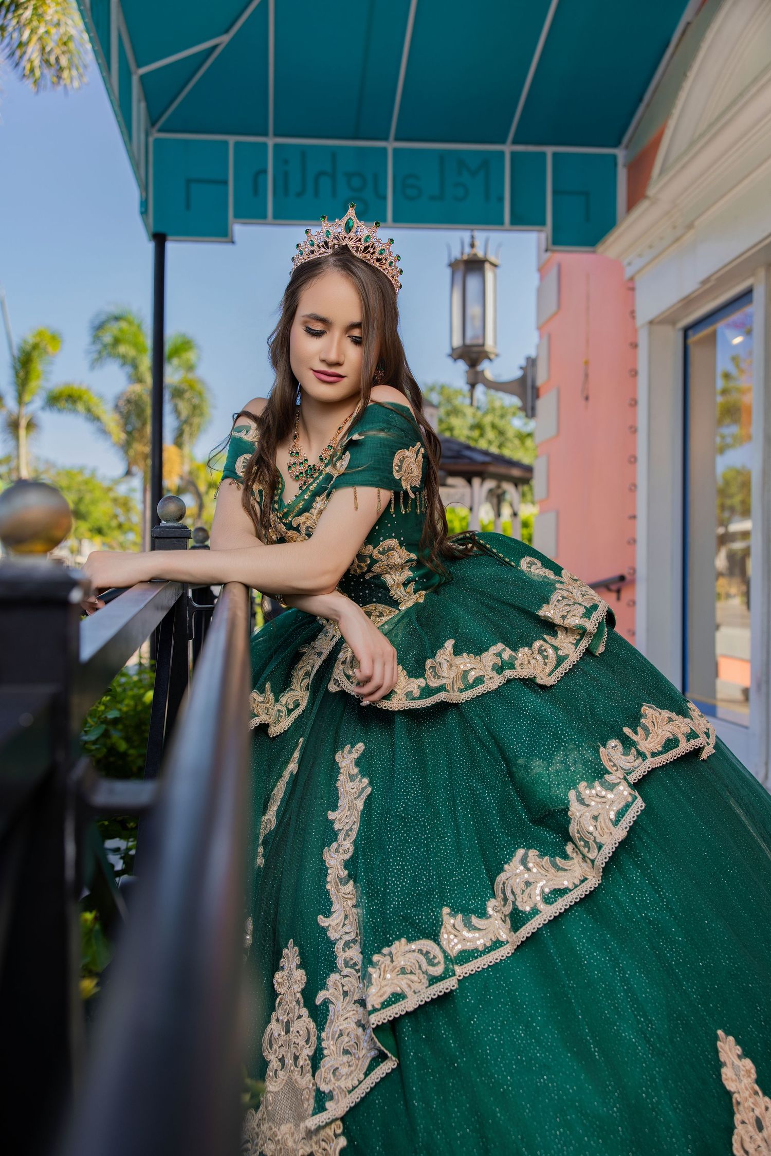 Young woman wearing an emerald green ballgown with gold lace embroidery posing on a balcony