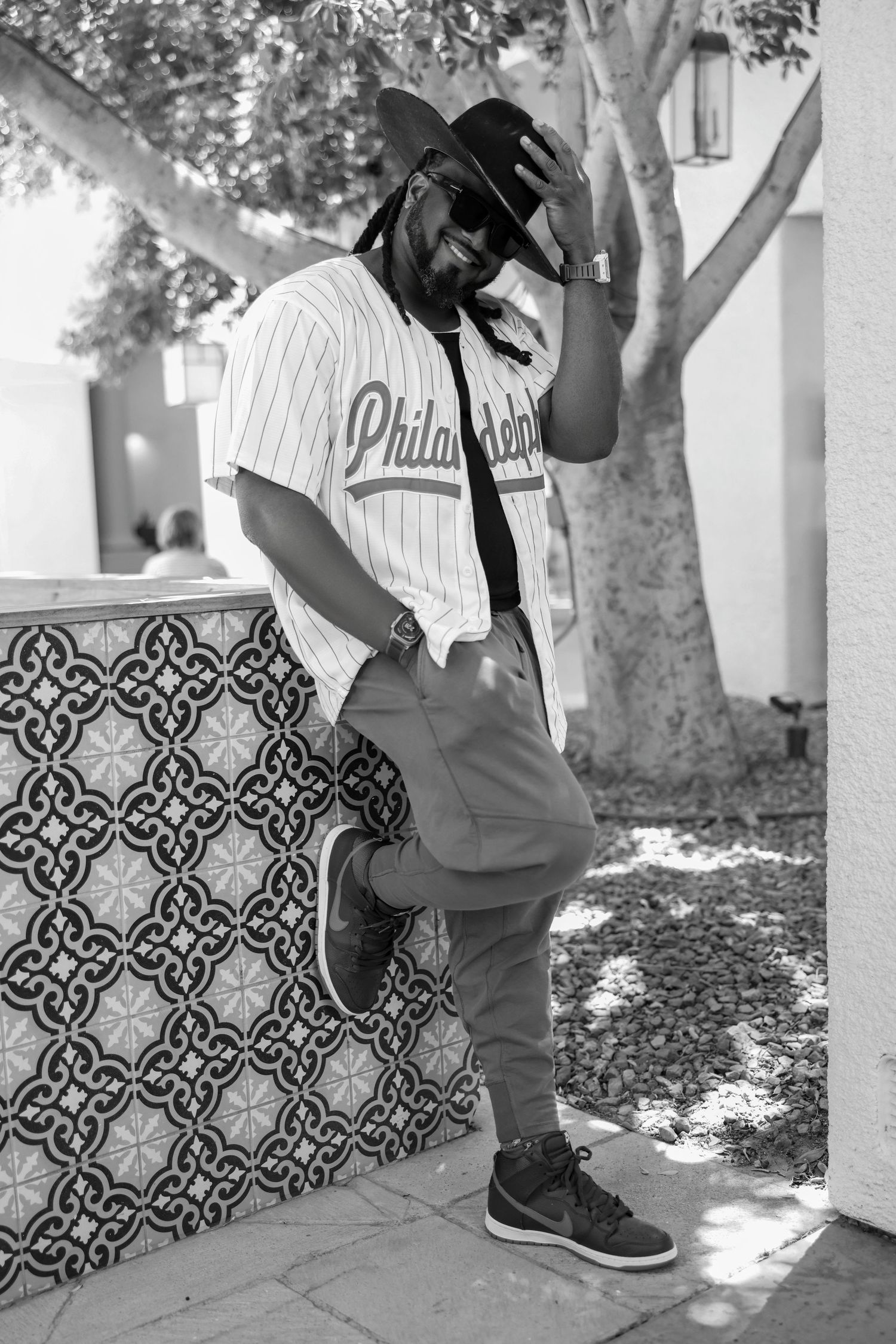 Black and white portrait of a stylish man in a Philadelphia jersey, brimmed hat, and sneakers leaning against a decorative tiled wall.