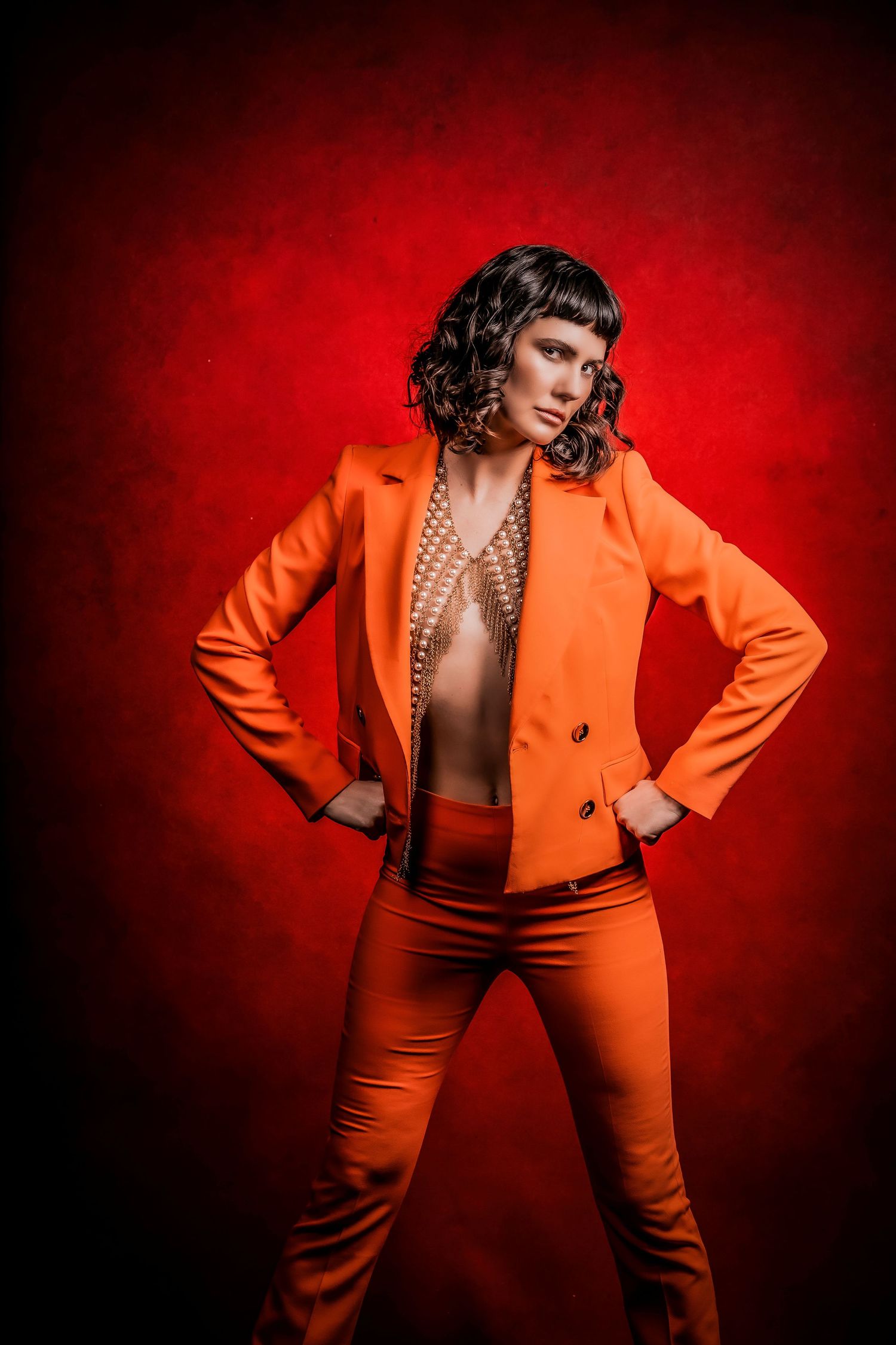 Woman with short dark hair wearing a vibrant orange pantsuit and layered gold body chains against a textured red background