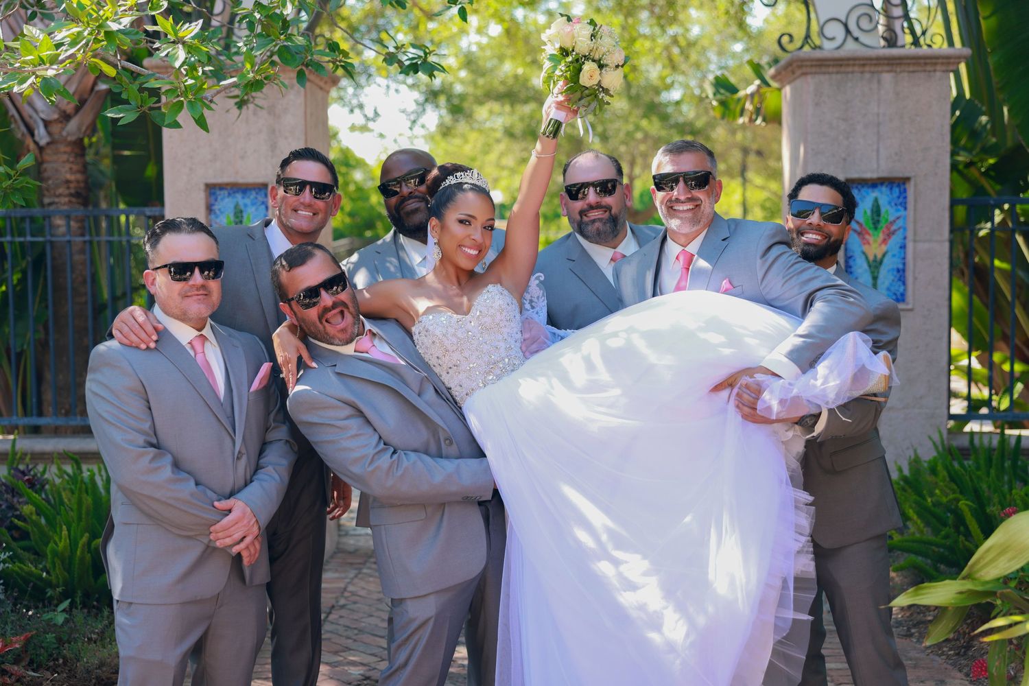 Groomsmen in gray suits playfully lifting and carrying the groom outdoors on a sunny wedding day