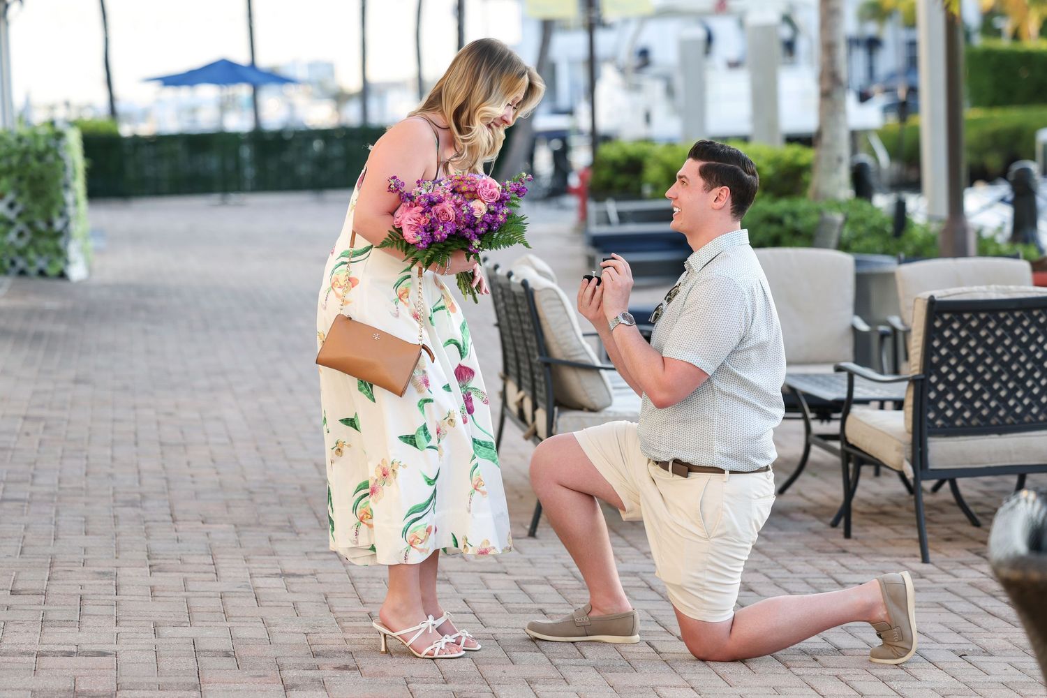 Man kneeling on an outdoor brick patio to propose with an engagement ring to a woman holding flowers