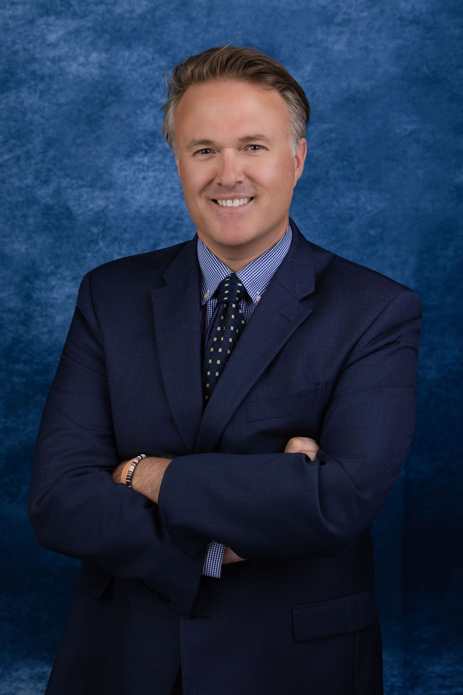 Smiling businessman with folded arms wearing a navy blue suit and patterned tie against a blue background