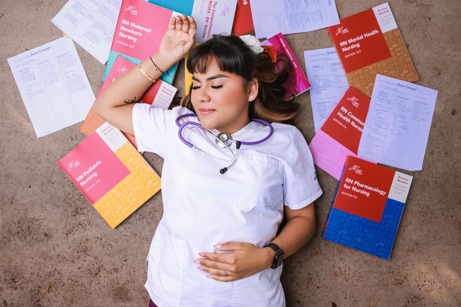 Overhead view of a graduate lying down surrounded by colorful textbooks, notebooks, and folders
