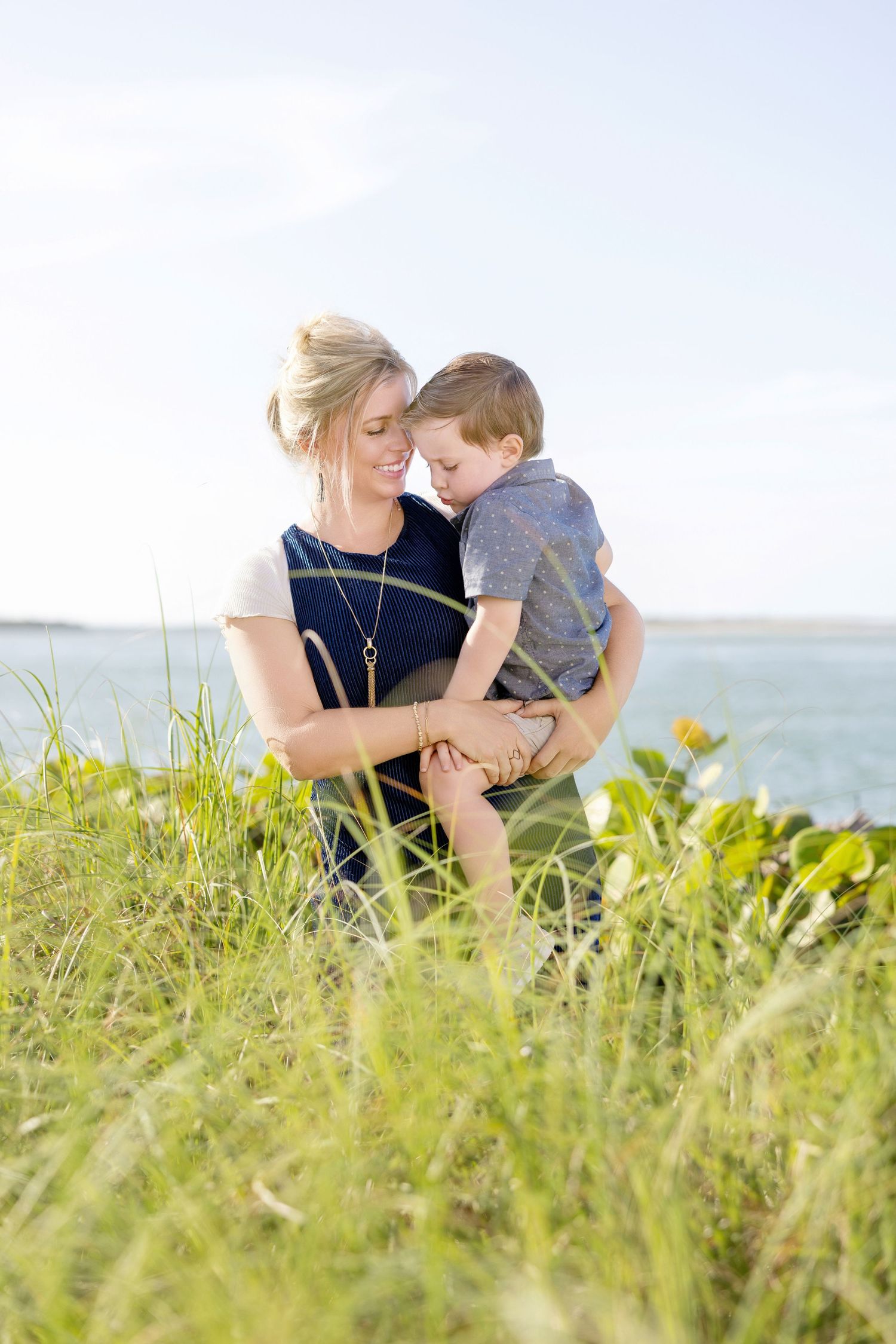 Mother holding her young son outdoors next to green coastal vegetation with an ocean view backdrop