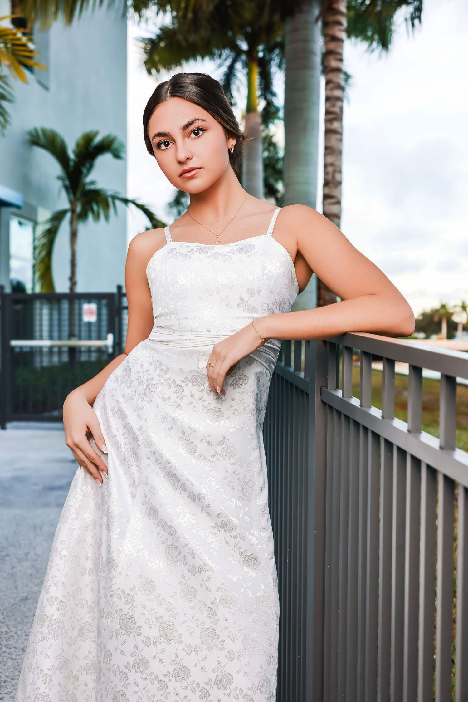 Young woman wearing a white floral lace dress leaning against a metal railing outdoors