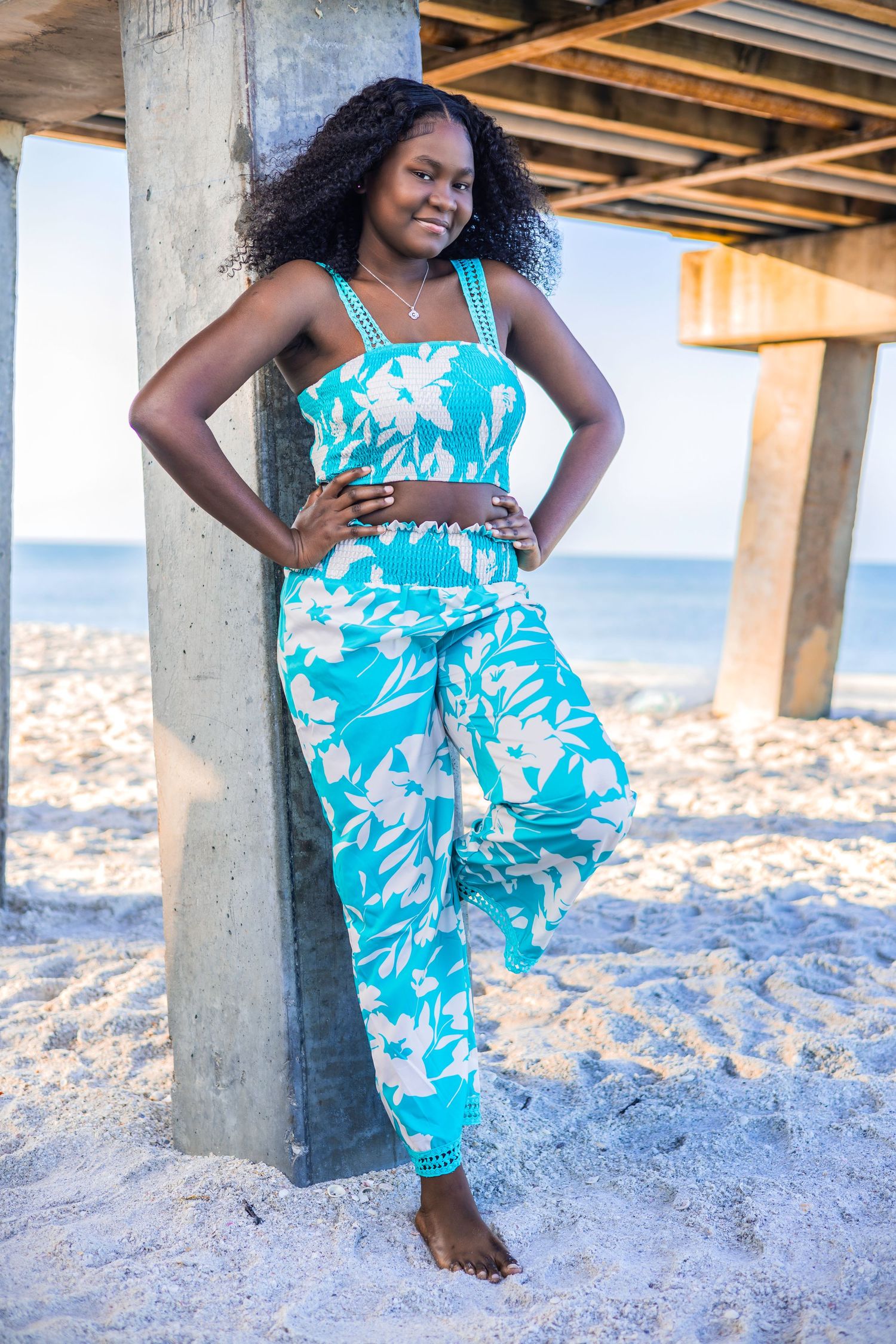 Smiling woman wearing a blue floral two-piece set leaning against a wooden beach pier pillar
