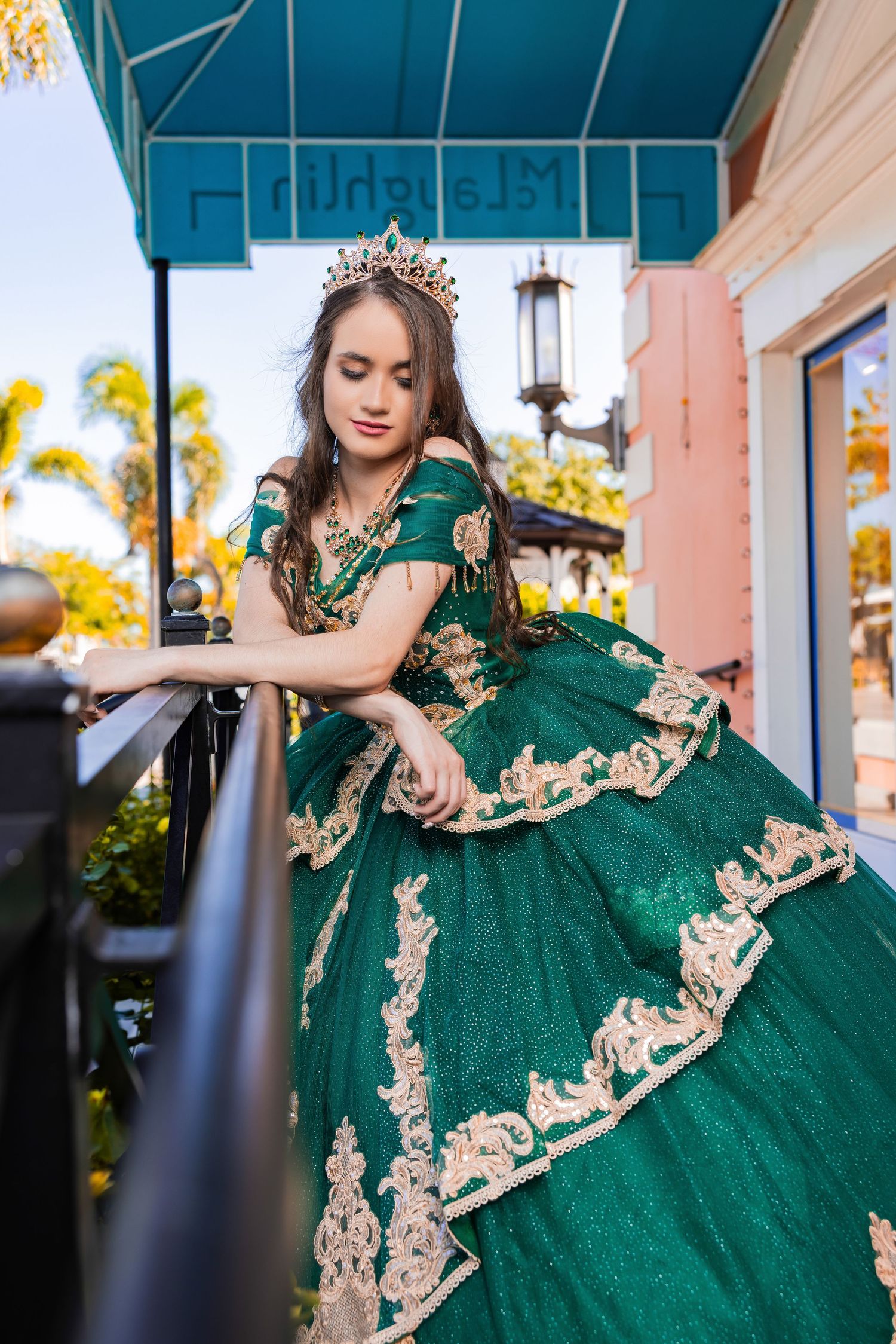 Quinceañera wearing an ornate emerald green ballgown with gold lace embroidery and a matching crown on a balcony