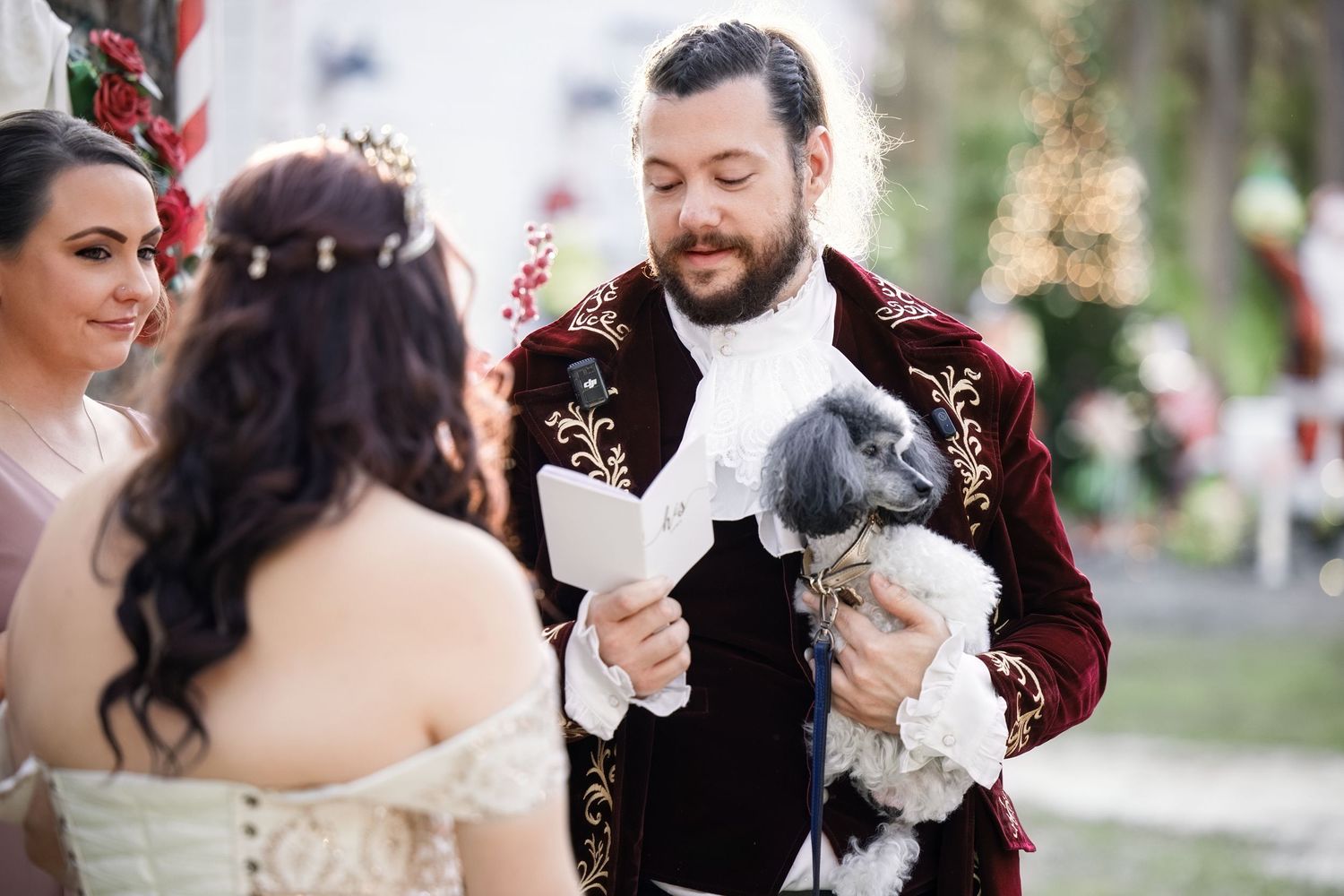 Groom reading wedding vows while holding a small dog next to his bride during an outdoor ceremony