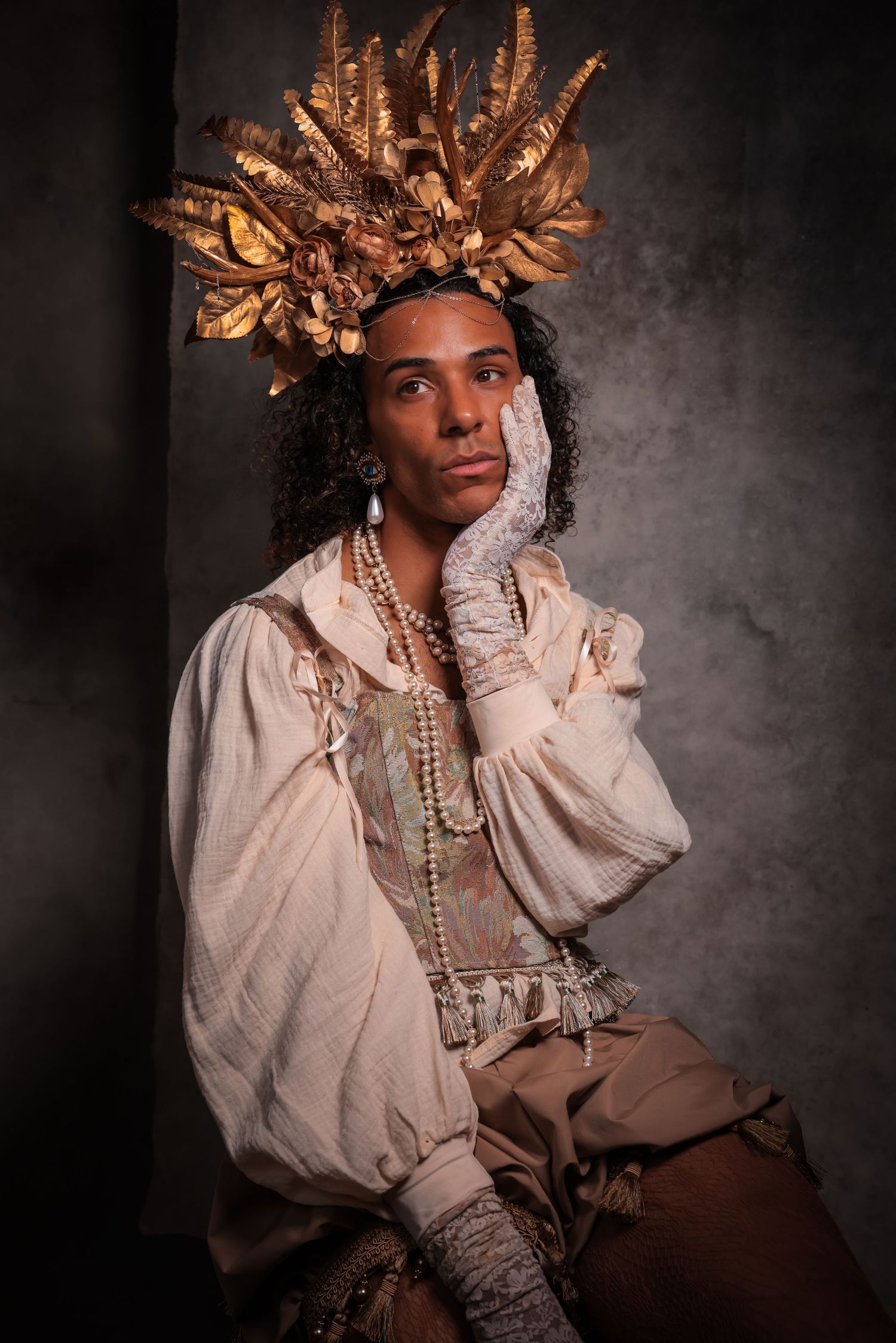 Pensive person in an ornate gold leaf crown and period lace clothing posing for a studio portrait