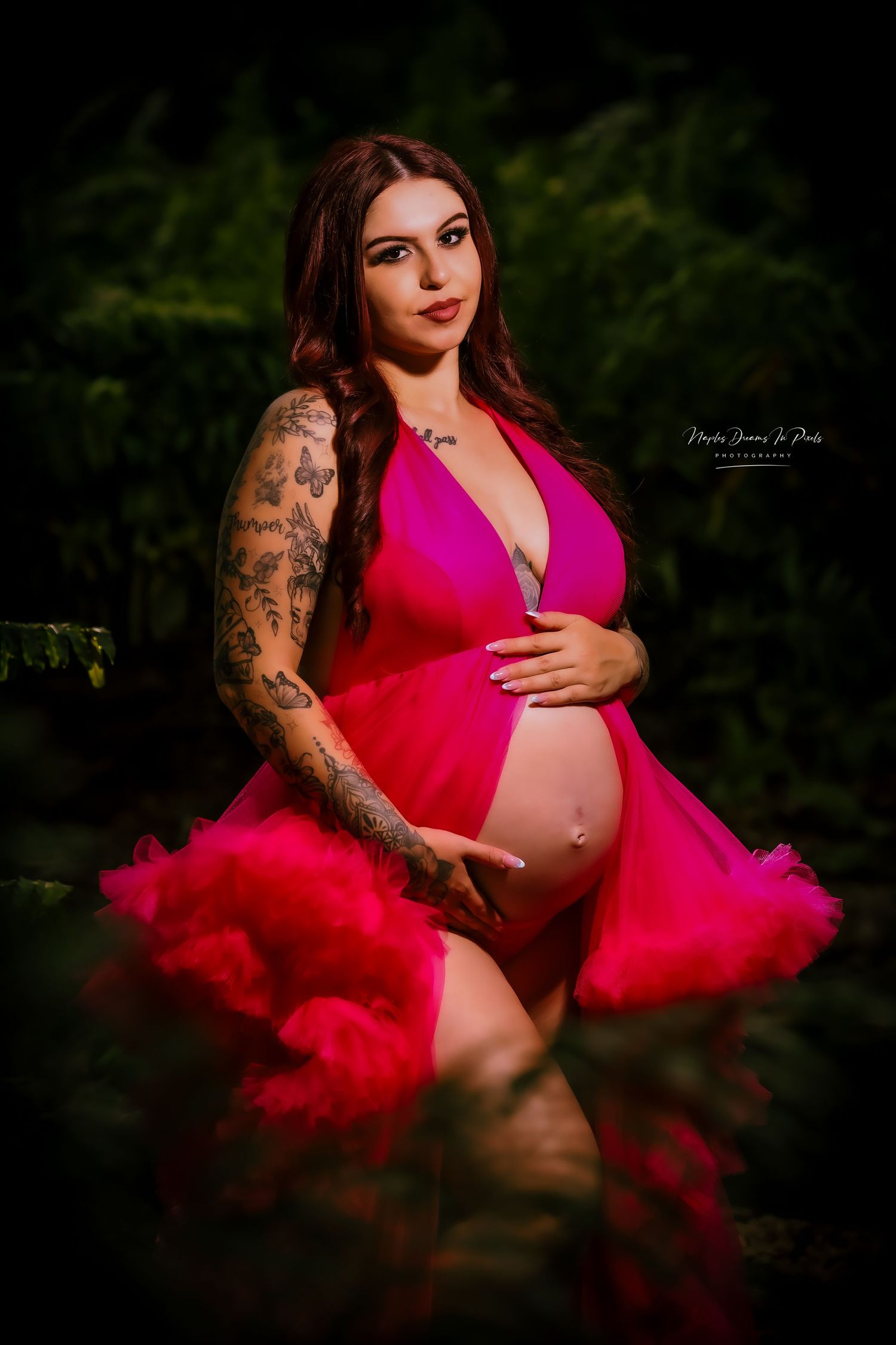 Dramatic portrait of a tattooed pregnant woman cradling her baby bump in an open hot pink maternity gown against a dark, moody background.