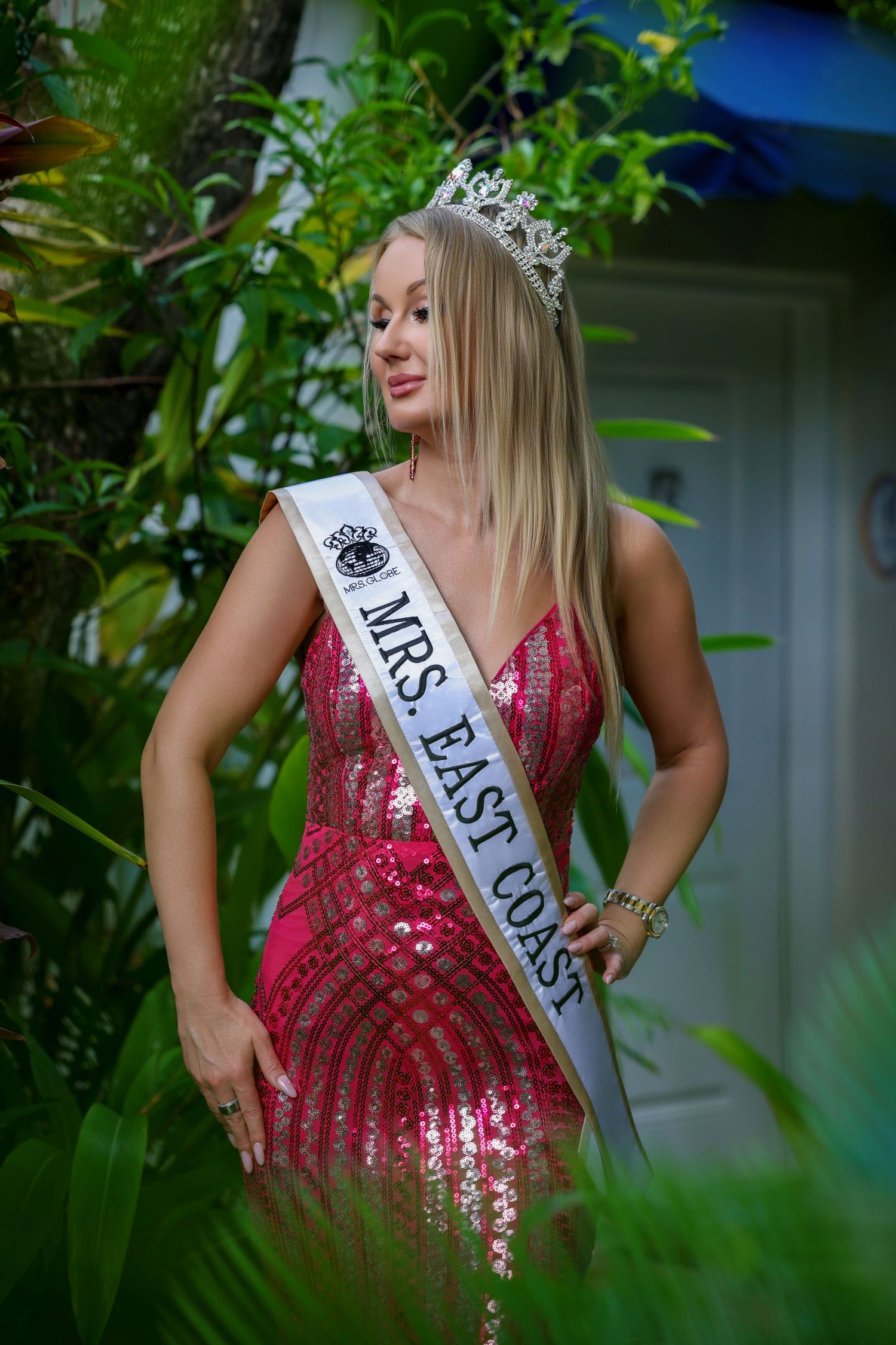 Pageant contestant wearing a pink sequined gown, crown, and Mrs. East Coast sash in a garden