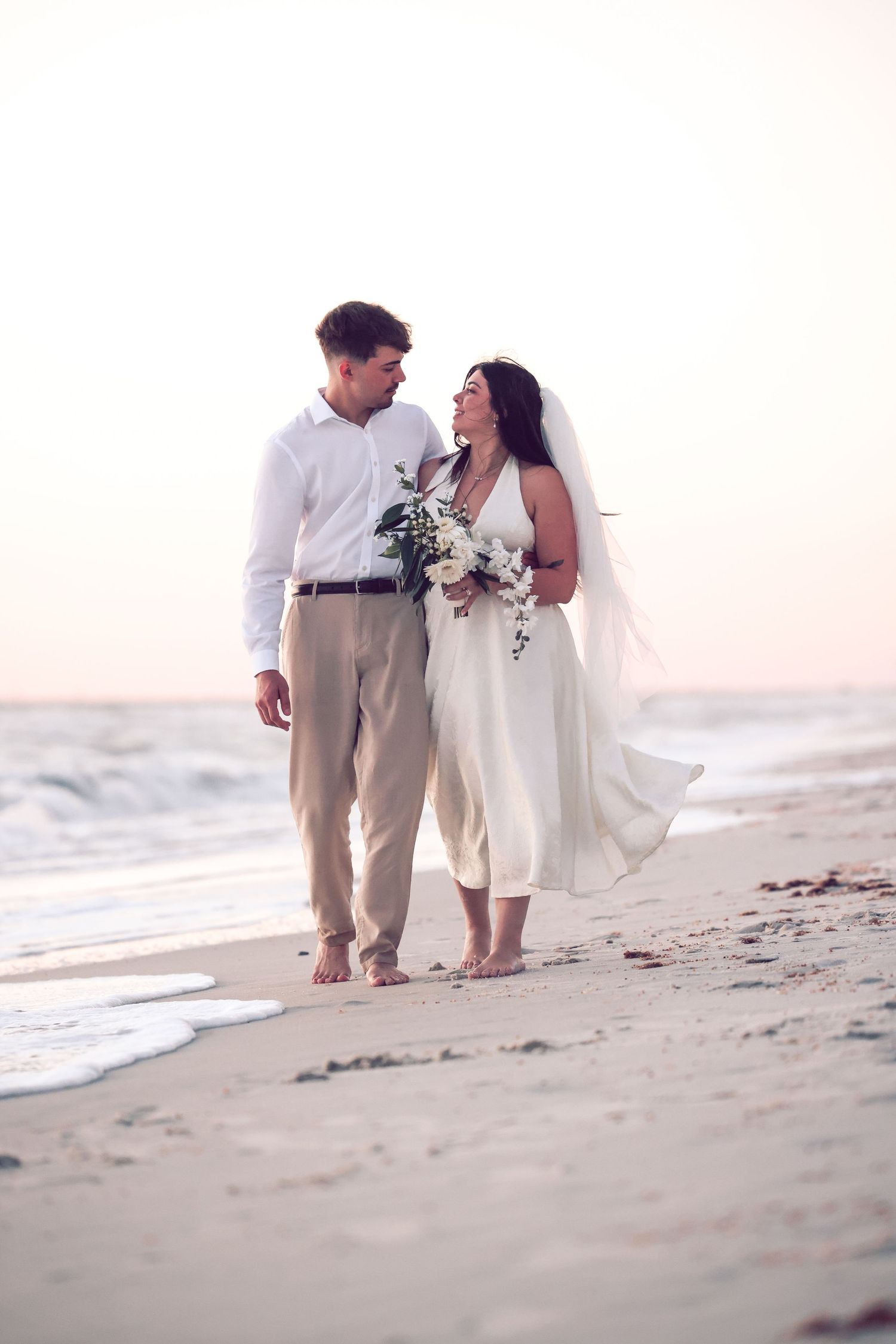 Newlyweds walking hand-in-hand along the ocean water line during a beach wedding at sunset