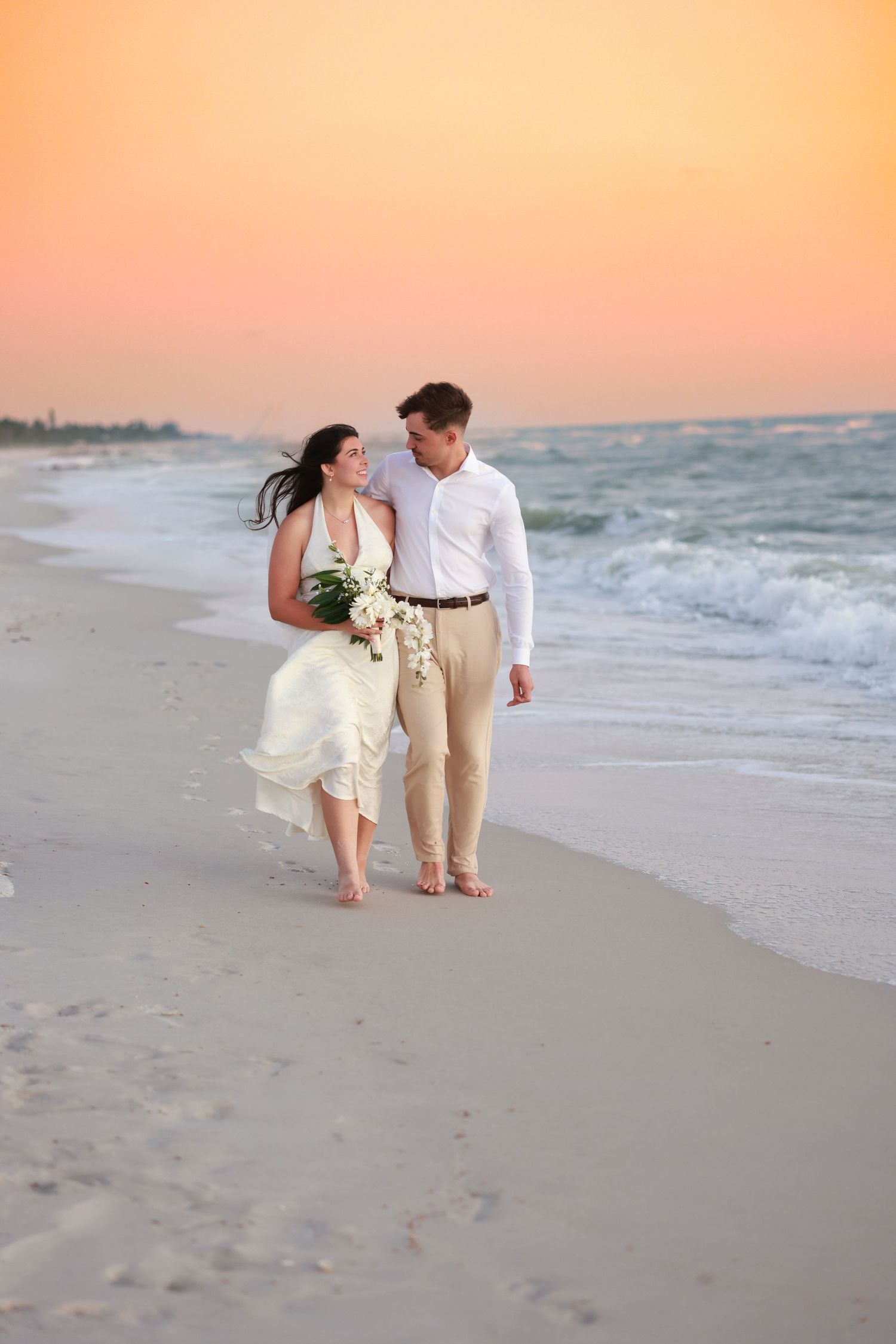 Bride and groom walking barefoot along the shoreline during a beach wedding at sunset