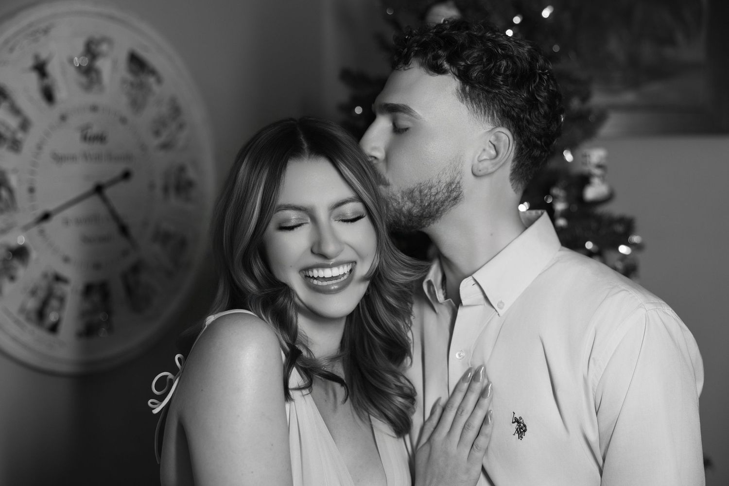 Black and white portrait of a man kissing a laughing woman on the temple in front of a clock wall decoration