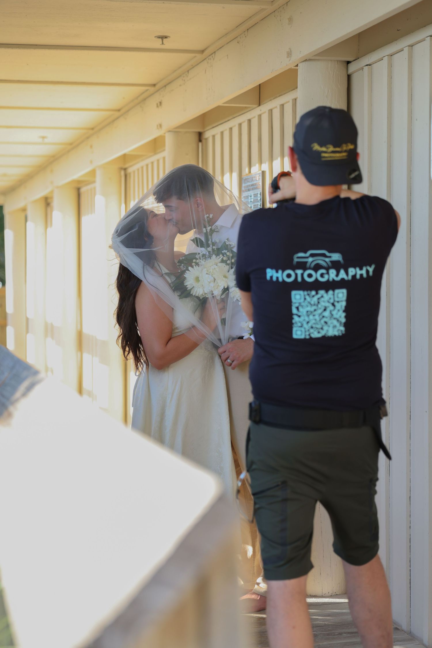 A professional wedding photographer wearing a black t-shirt captures a candid moment of a bride and groom kissing in a bright hallway.