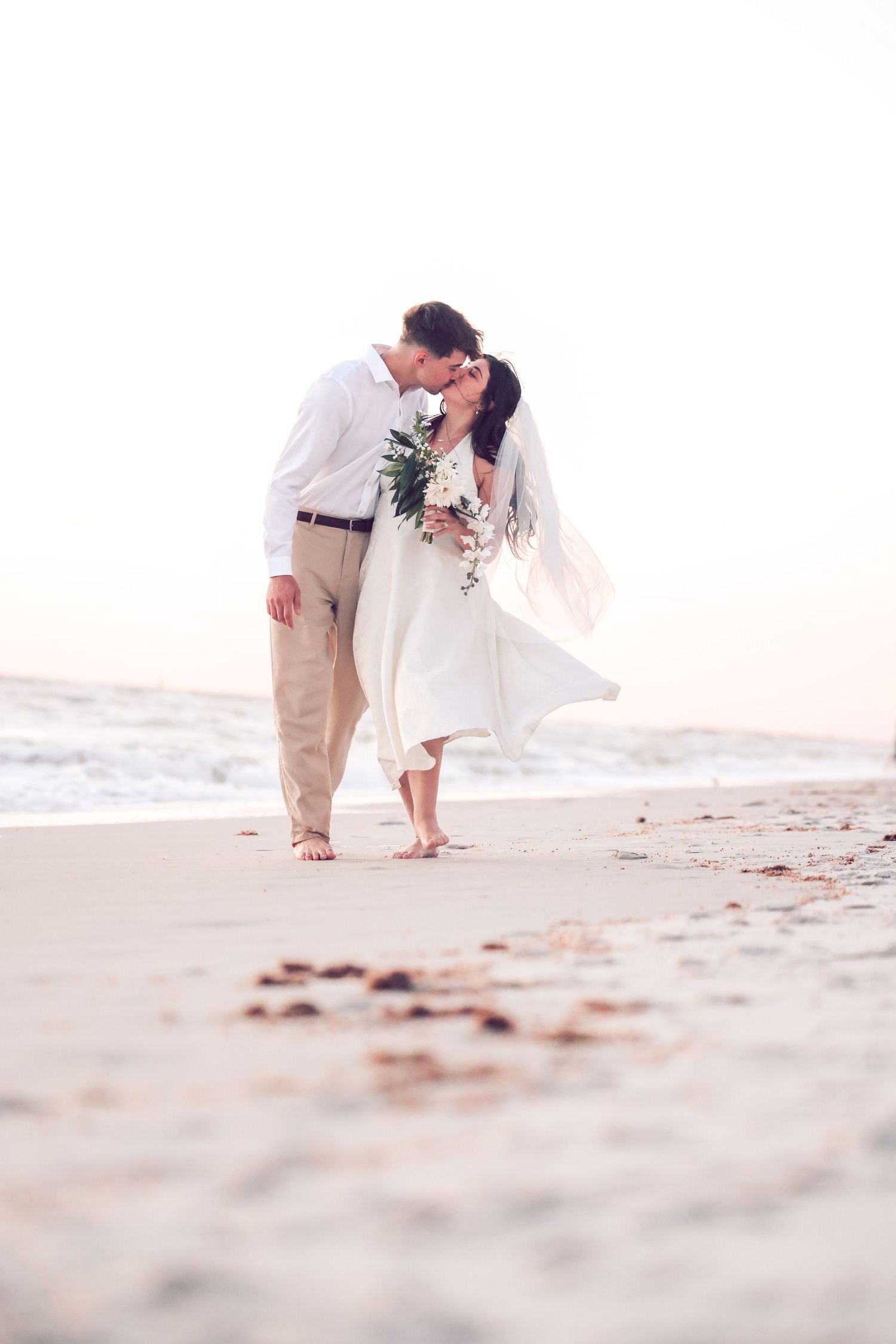 Bride and groom kissing barefoot on a sandy beach shore during an outdoor wedding ceremony