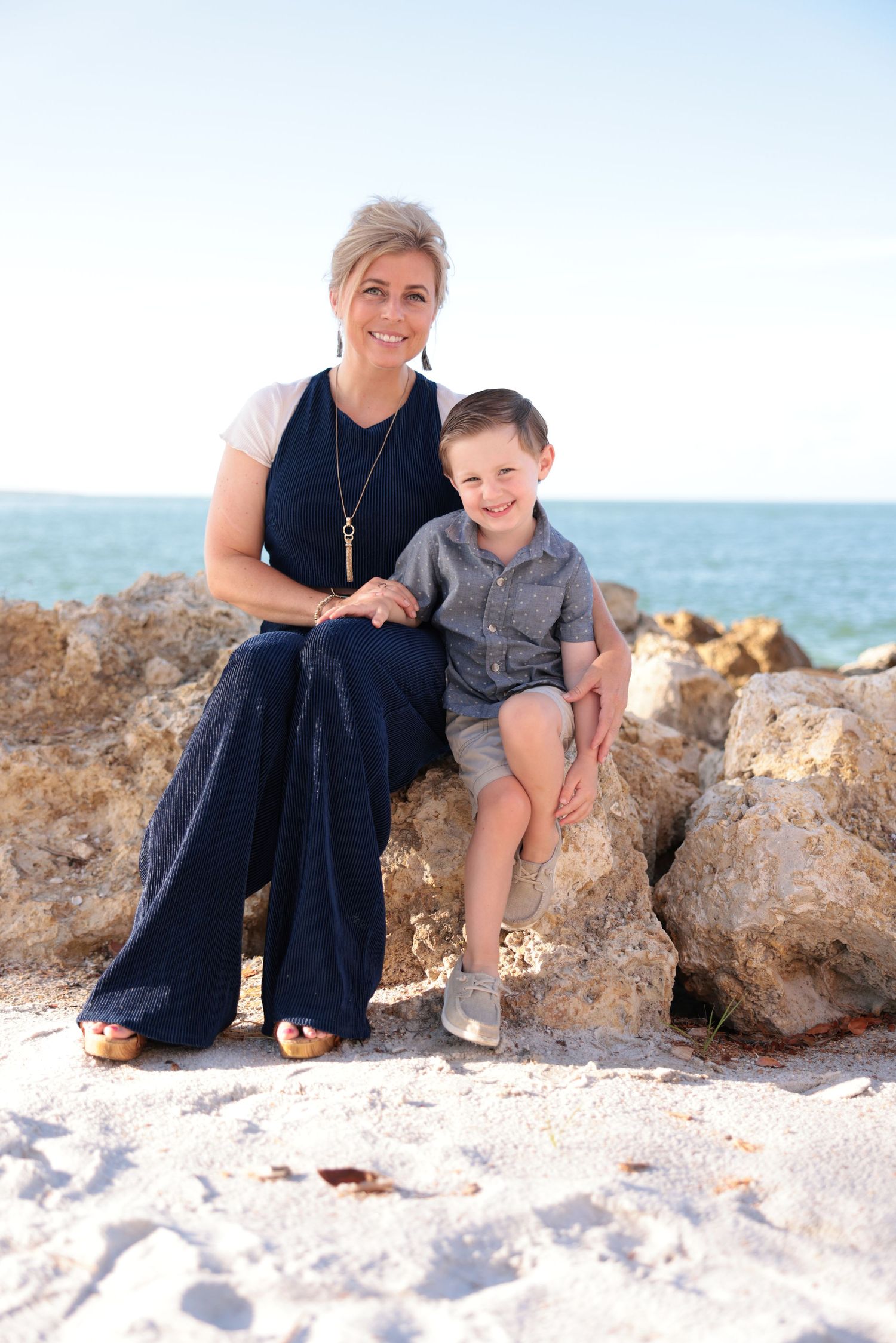 Mother and young son smiling while sitting together on beach rocks near the ocean.
