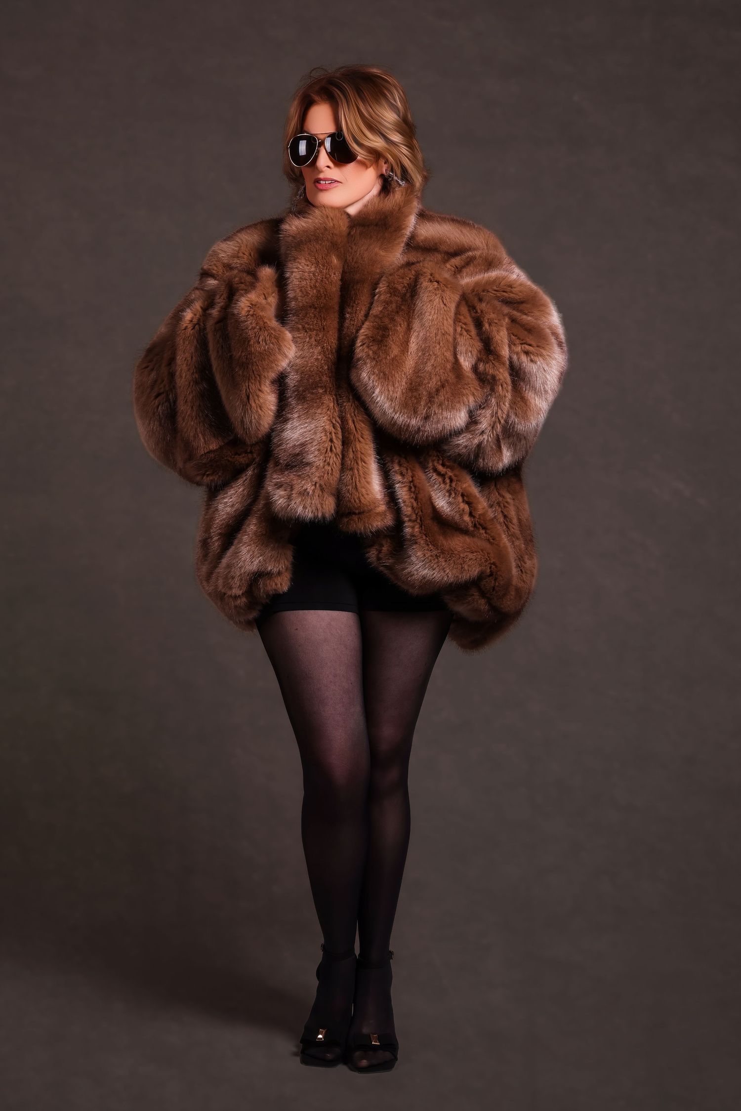 Full-length studio portrait of a woman wearing sunglasses and a voluminous, oversized brown fur coat with black tights.
