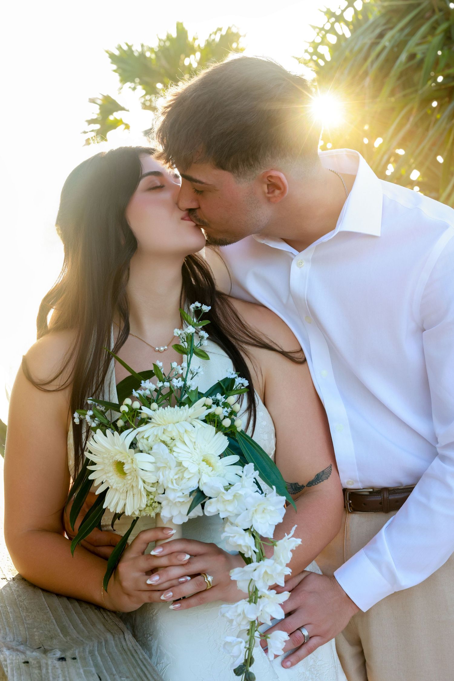 Bride holding a white floral bouquet while kissing her groom during a sunny outdoor wedding