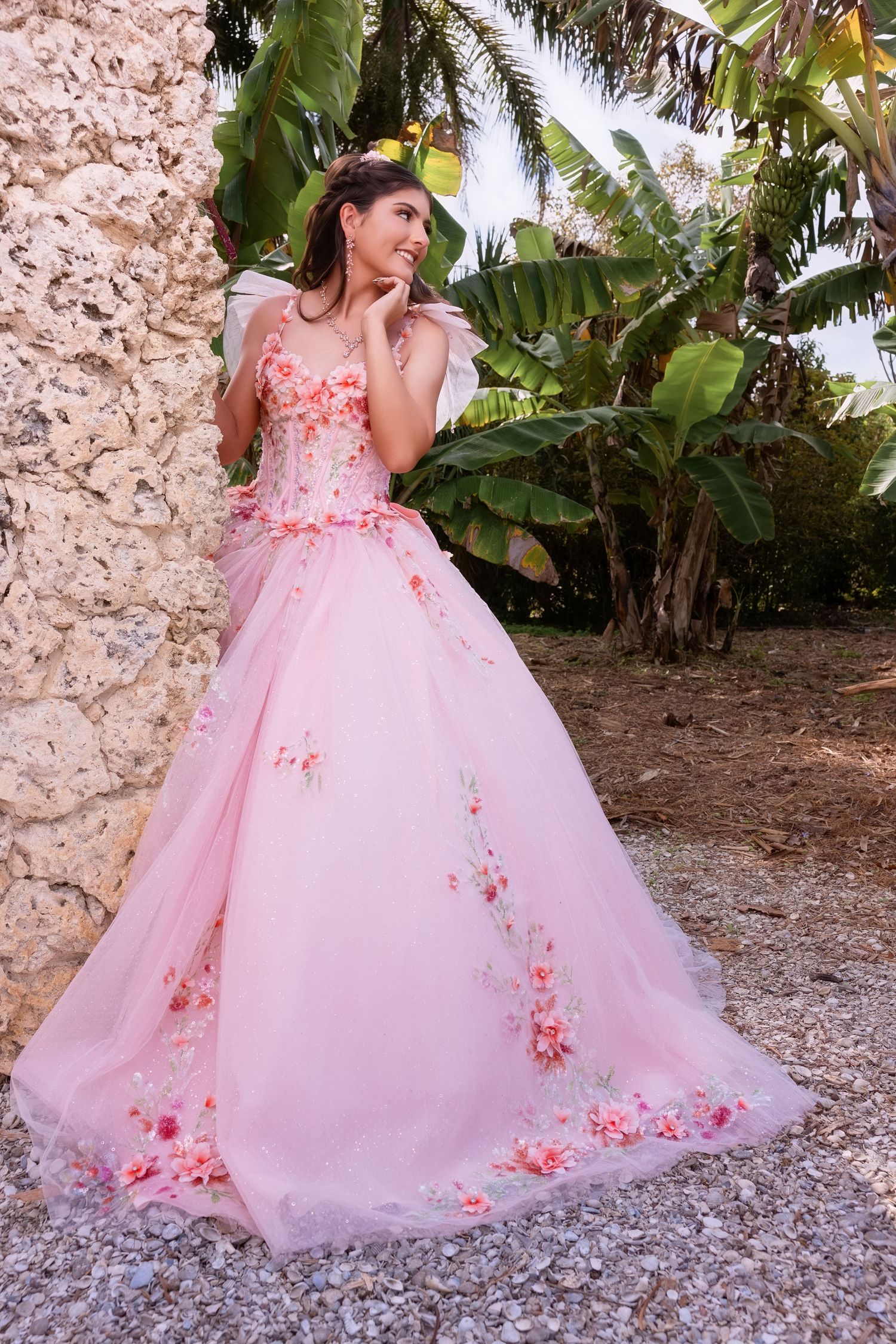 Young woman in an off-the-shoulder pink floral embroidered ballgown posing in a lush tropical garden