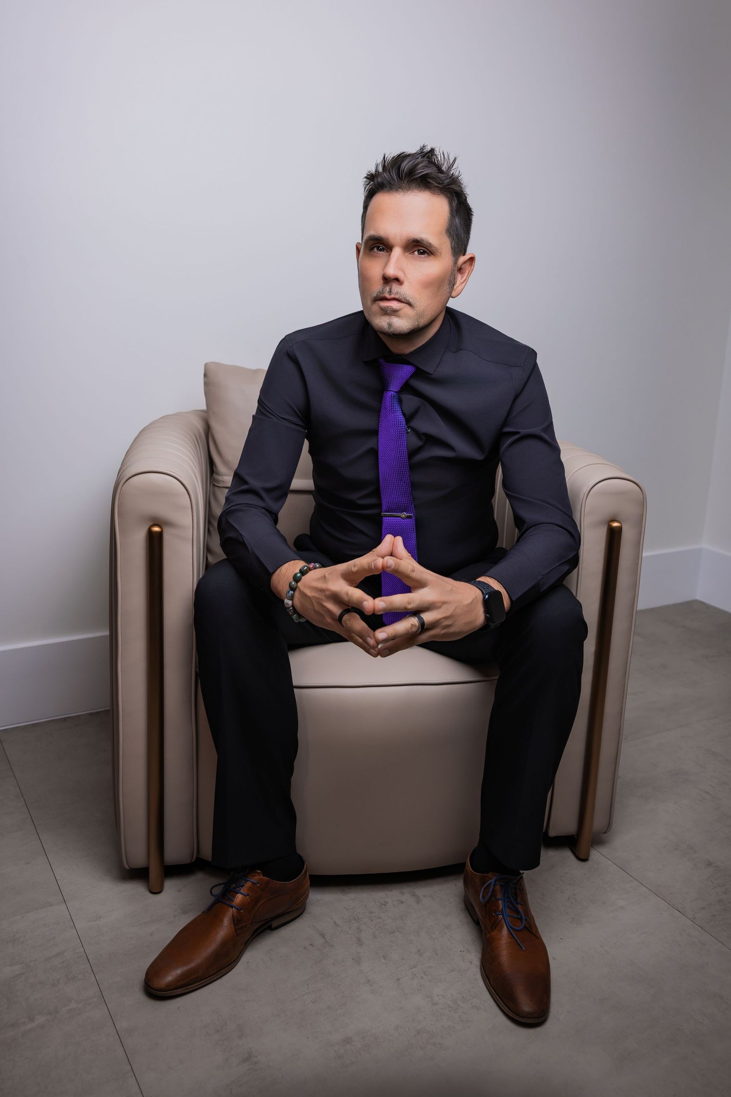 Man wearing a black tailored suit, bright purple tie, and brown dress shoes sitting in an armchair