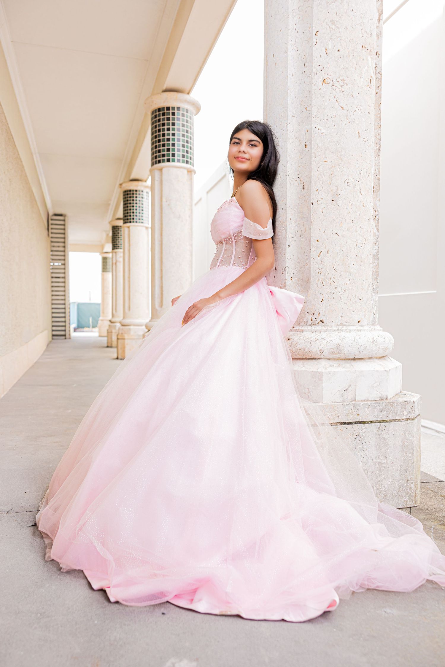 Young woman in a pink off-the-shoulder tulle ballgown leaning against a classical architectural pillar.