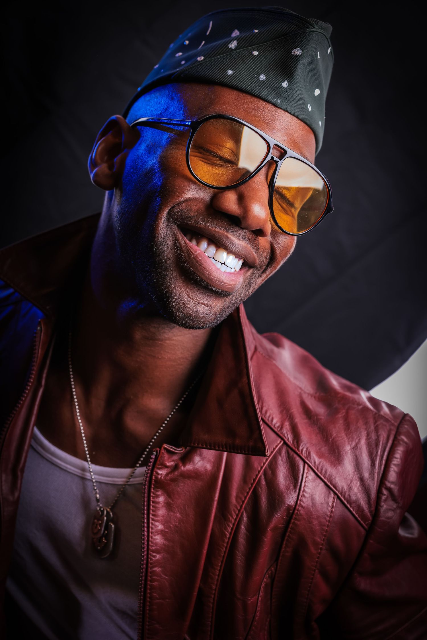 Smiling man wearing a brown leather jacket, tinted sunglasses, and a patterned cap in a studio portrait